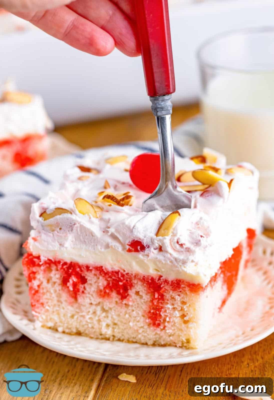 a fork shown being inserted by a hand into a slice of cherry almond poke cake that is on a white plate. 