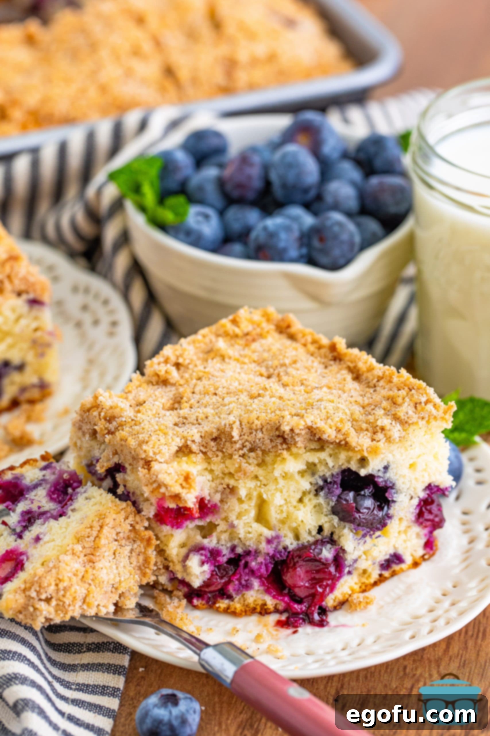 Sweet Blueberry Streusel Cake 10 A close-up of a slice of blueberry buckle on a white plate, with a fork gently scooping out a piece, highlighting the moist interior and crumbly topping.