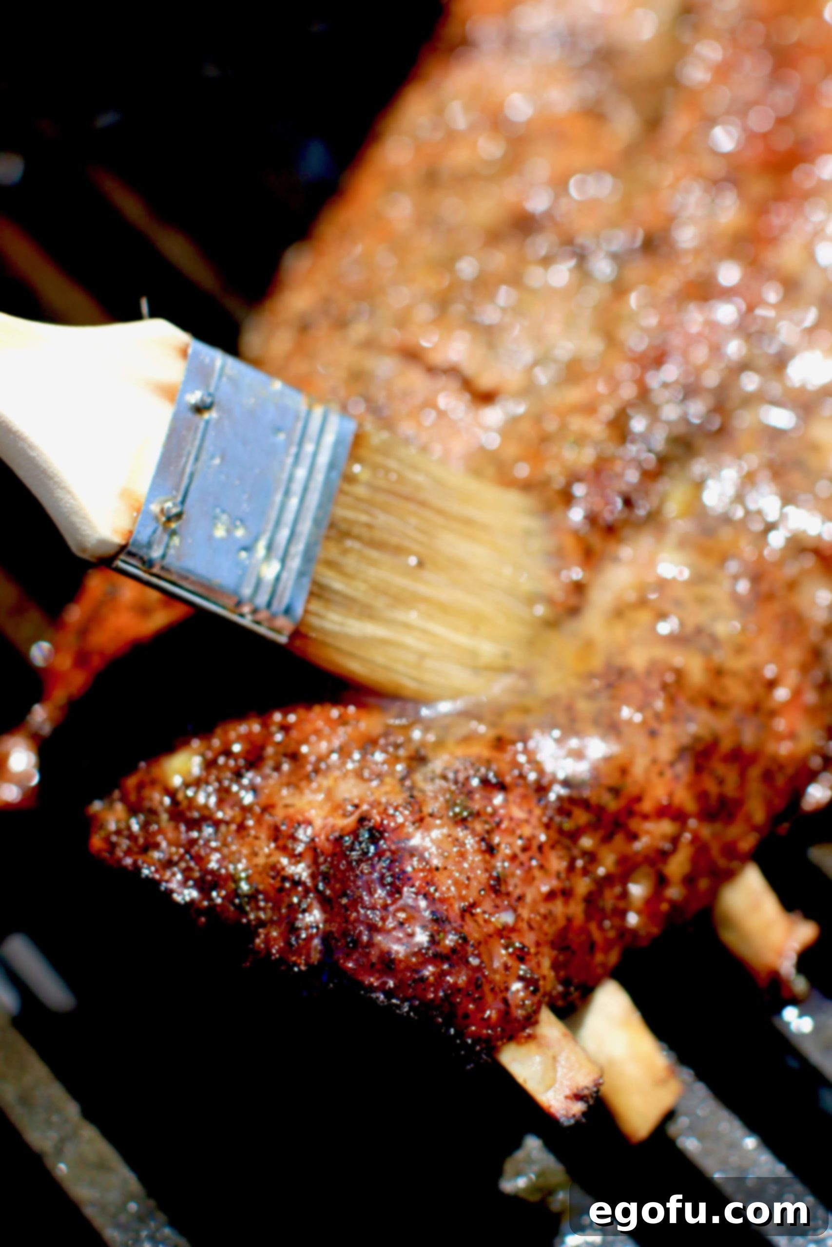 A basting brush applying a rich, shiny glaze onto hot pork ribs cooking on a gas grill.