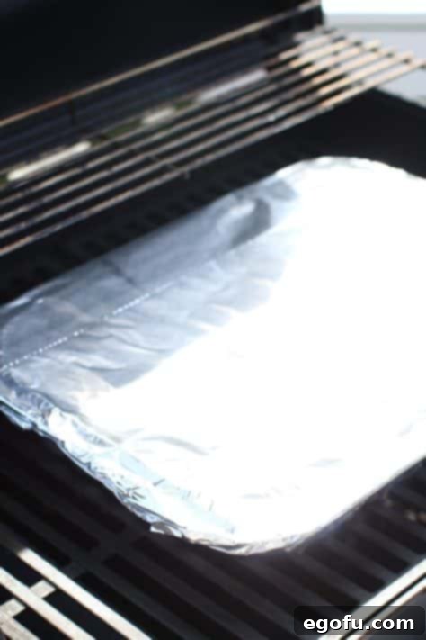 a covered aluminum foil pan showed on the grate of a gas grill.