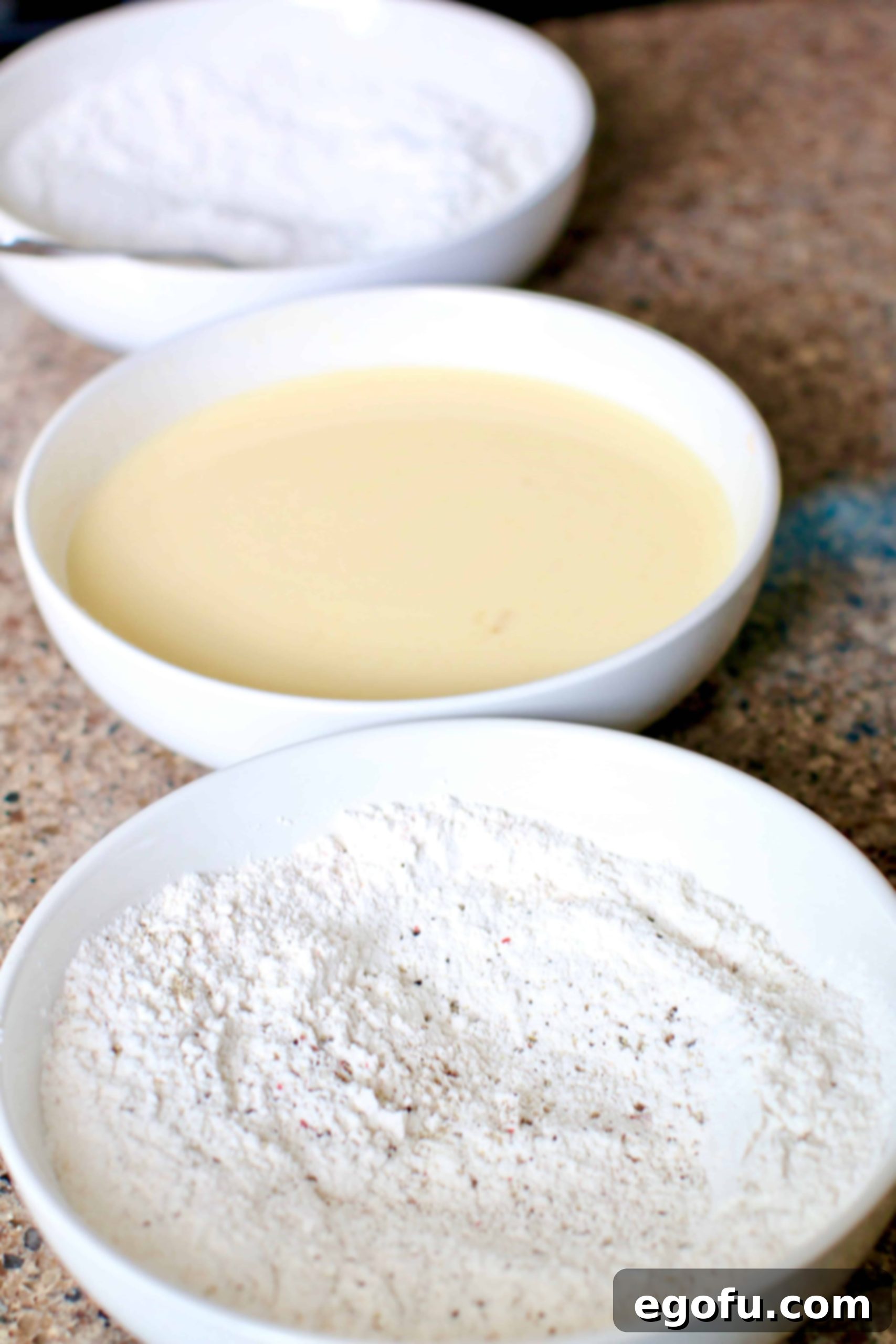 Three shallow bowls set up for fried chicken coating, containing seasoned all-purpose flour, buttermilk and eggs mixture, and seasoned flour.