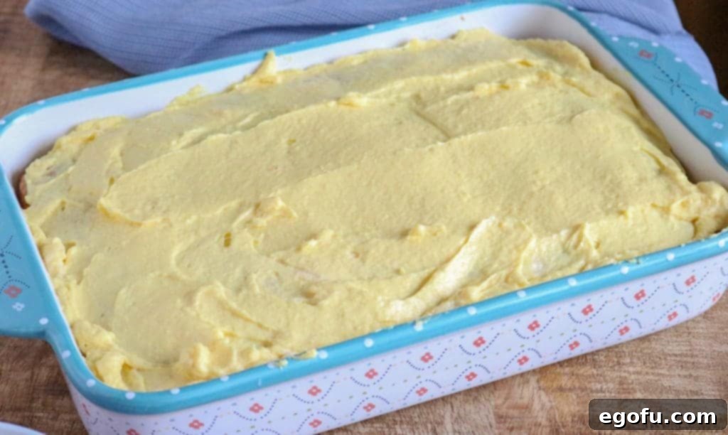 Creamy prepared vanilla pudding being poured evenly over the top of a baked cake that has been poked with holes, ensuring it soaks into every crevice.