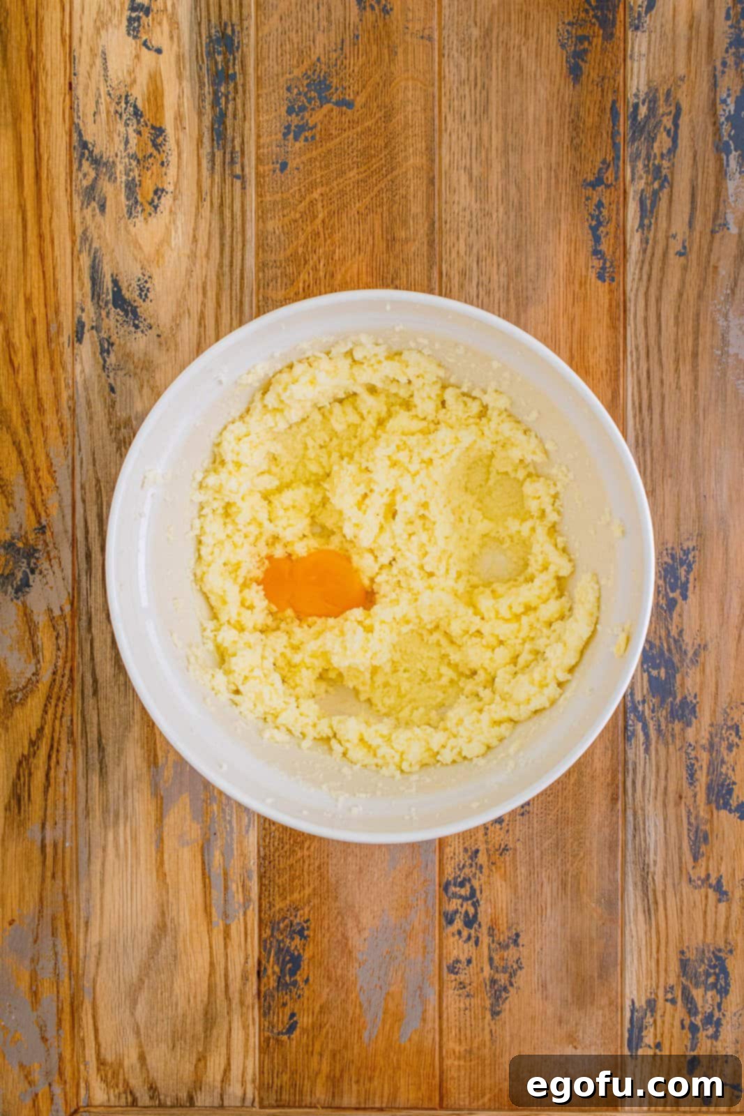 A hand adding egg yolks, one by one, to the creamed butter and sugar mixture in a large white bowl, ensuring thorough blending.