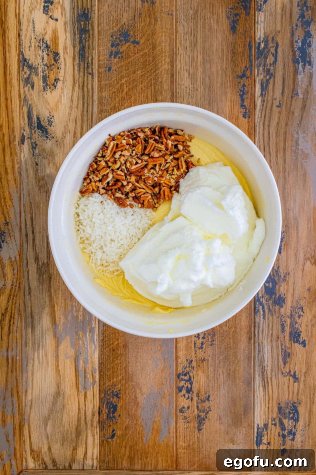A mix of egg whites, chopped pecans, and shredded sweetened coconut being gently folded into the Italian Cream Cake batter in a large white bowl.