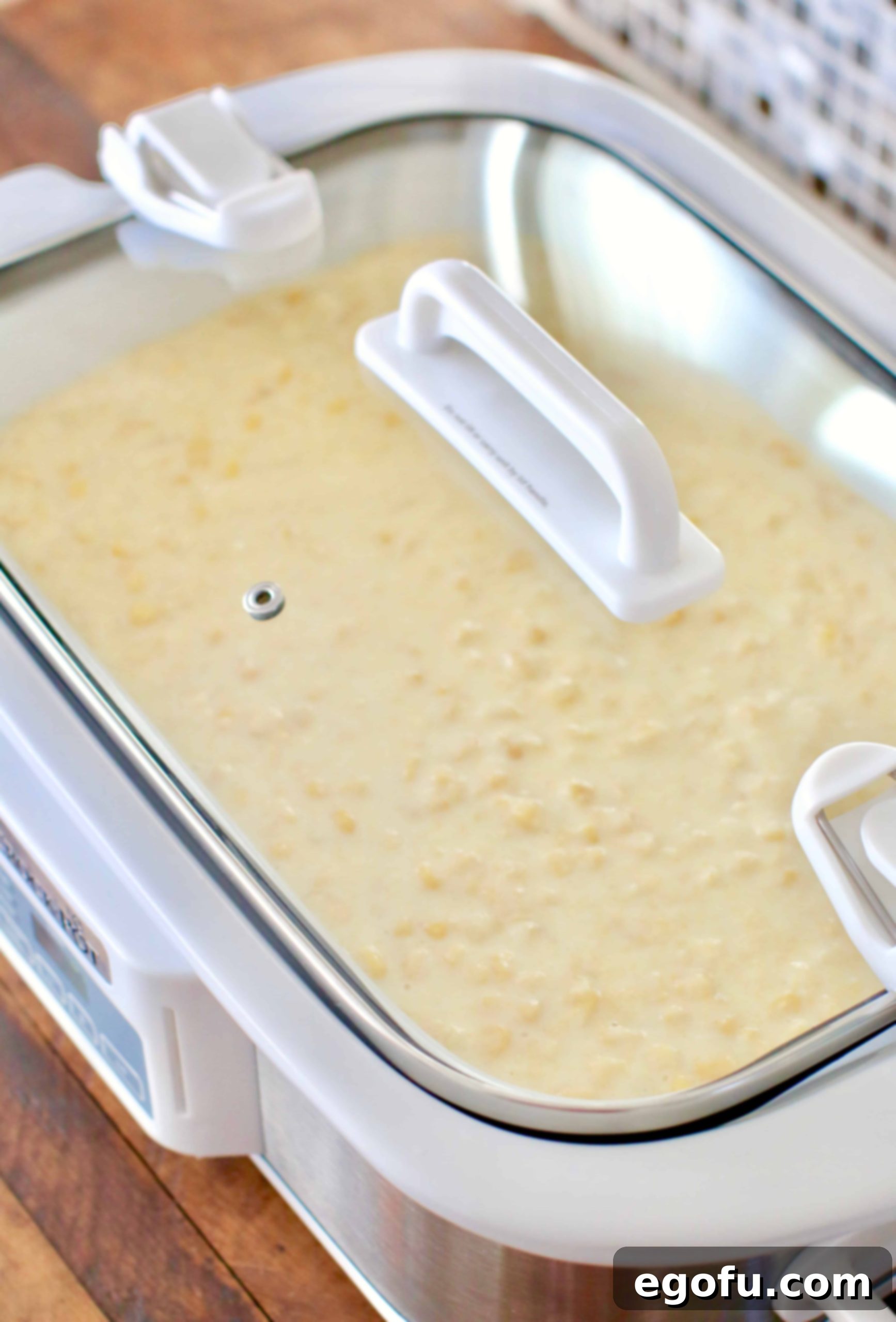 A casserole crock pot, covered with a clear lid, containing the uncooked corn casserole batter, set to begin the slow cooking process.