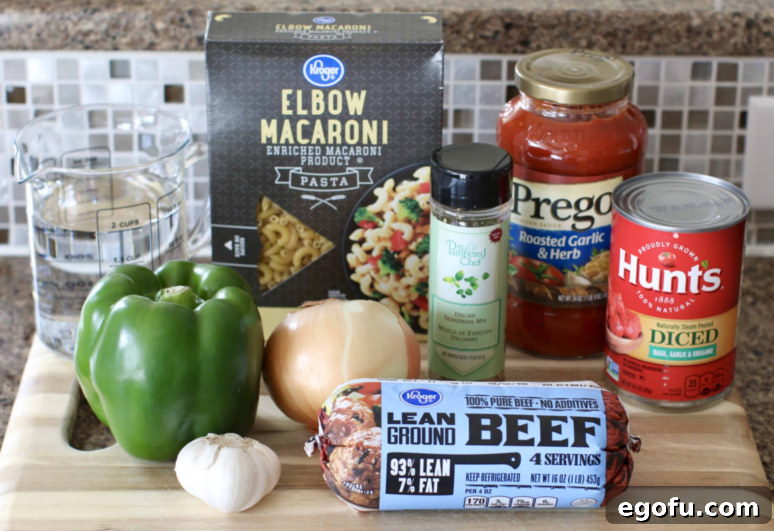 Hearty Instant Pot American Goulash 5 A flat lay arrangement of all ingredients for Instant Pot American Goulash: lean ground beef, a small yellow onion, green bell pepper, fresh garlic cloves, a can of diced tomatoes, a jar of spaghetti sauce, a box of macaroni noodles, a bottle of Italian seasoning, and a bag of shredded cheddar cheese.
