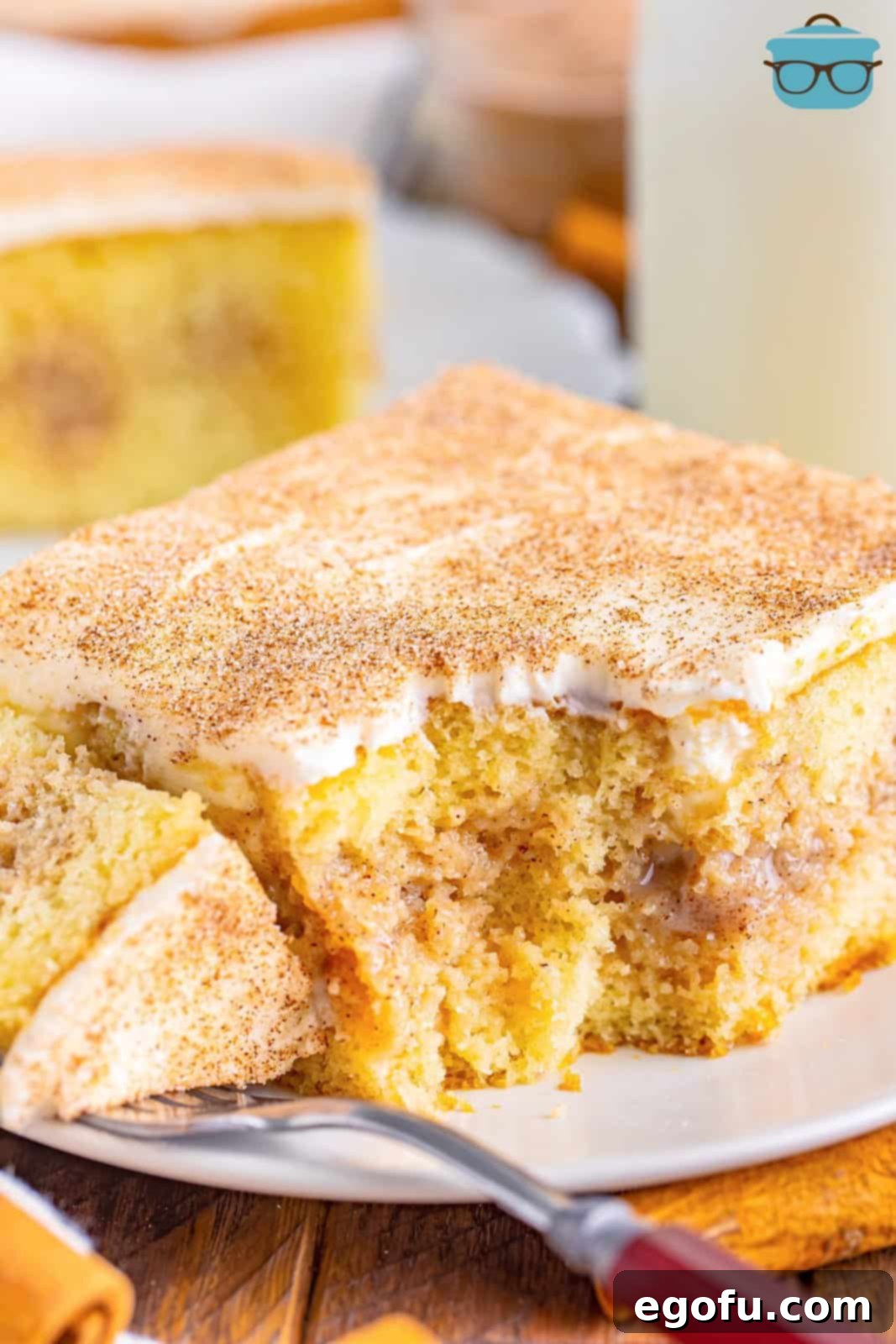 Cinnamon Swirl Poke Cake 2 A perfectly sliced piece of Cinnamon Roll Poke Cake on a small white plate, showing the moist cake, rich filling, and creamy frosting, with a fork holding another bite.