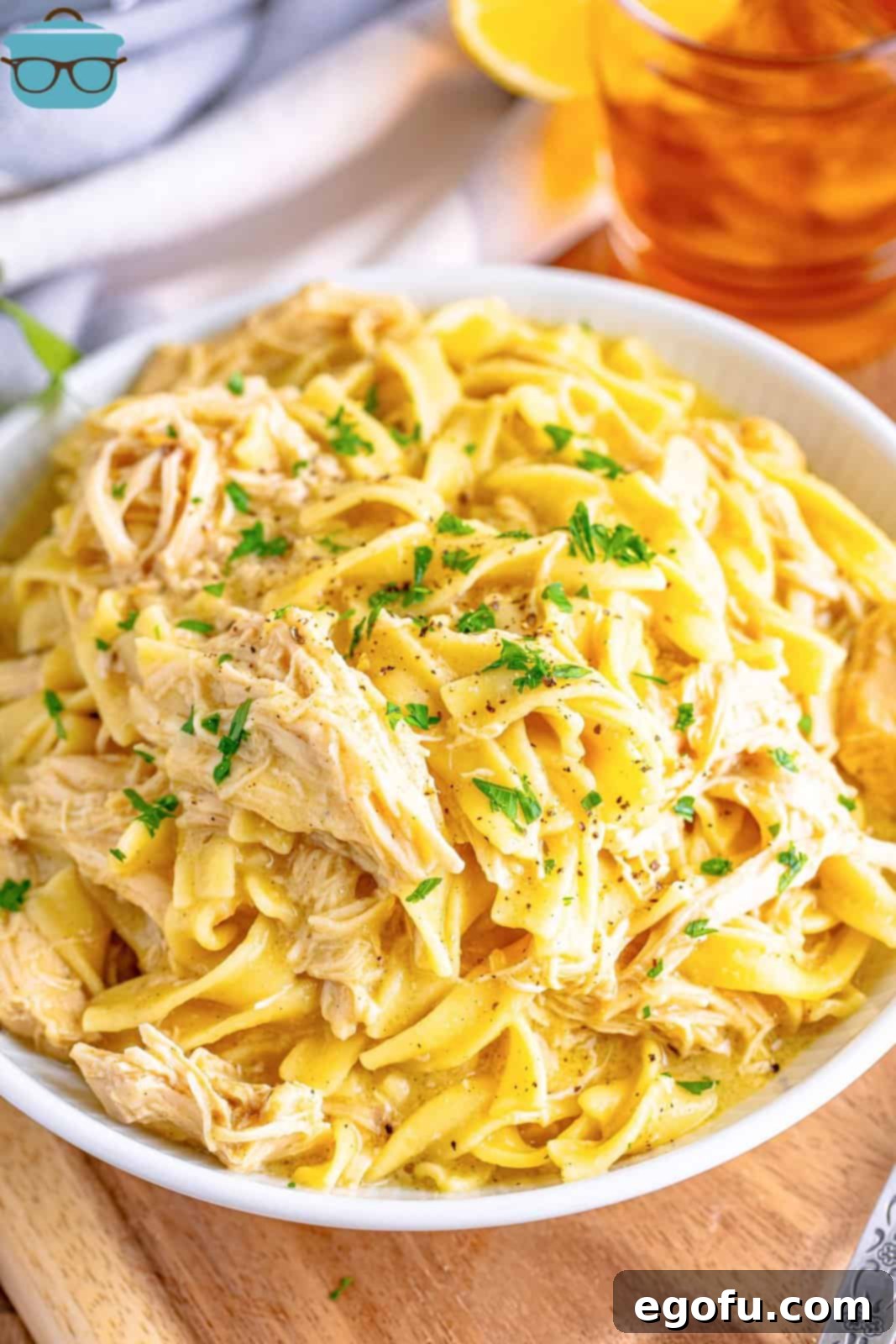 Closeup view of a large white serving bowl filled with creamy crock pot chicken and noodles, with a glass of iced tea with lemon in the background, highlighting its comforting appeal.