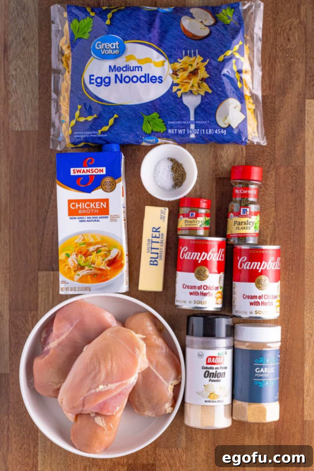 All ingredients for Crock Pot Chicken and Noodles laid out: boneless, skinless chicken breasts, cans of cream of chicken soup, various spices (salt, pepper, poultry seasoning, garlic powder, onion powder), salted butter, chicken broth, and medium egg noodles, ready for assembly.