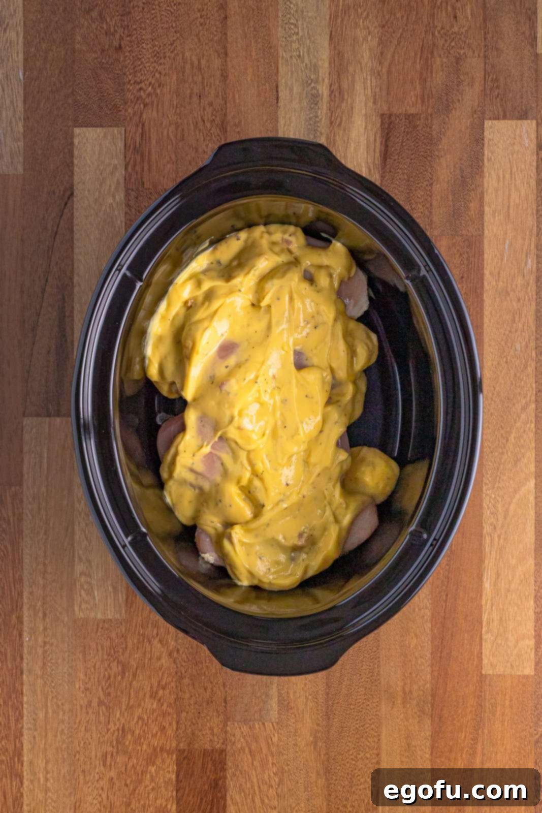 Chicken breasts covered with cream of chicken soup in an oval black slow cooker, ready for the next layer of ingredients.