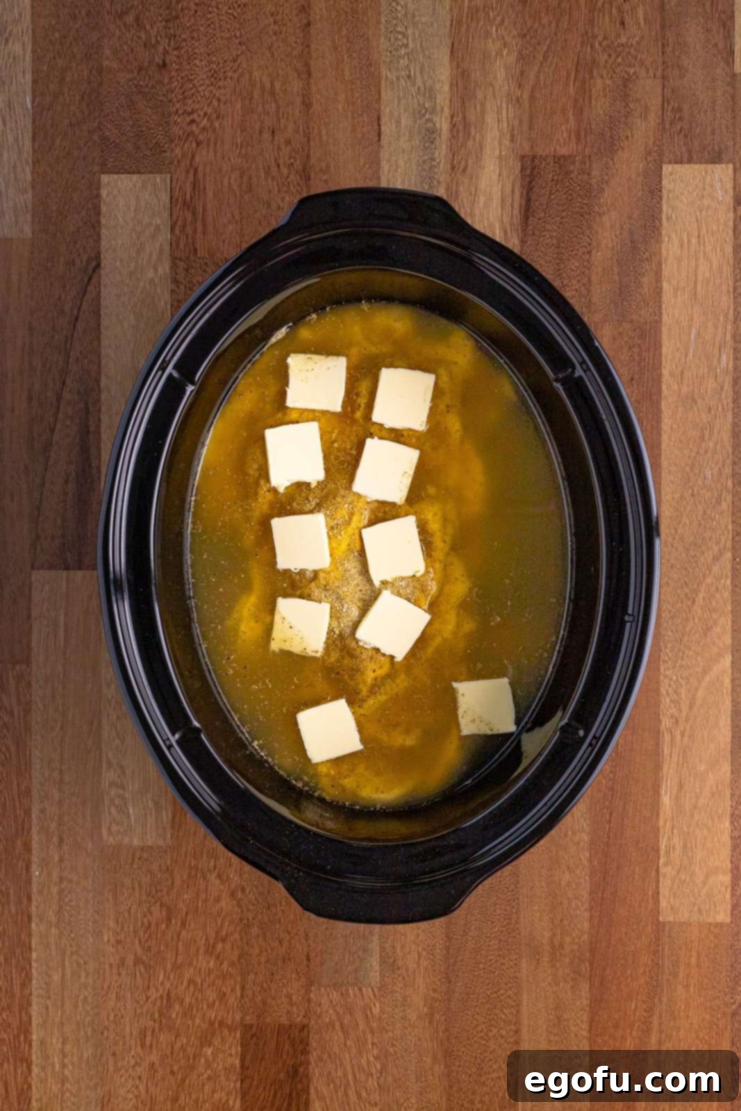 Chicken, cream of chicken soup, spices, and butter slices submerged in chicken broth in an oval slow cooker, ready for cooking.