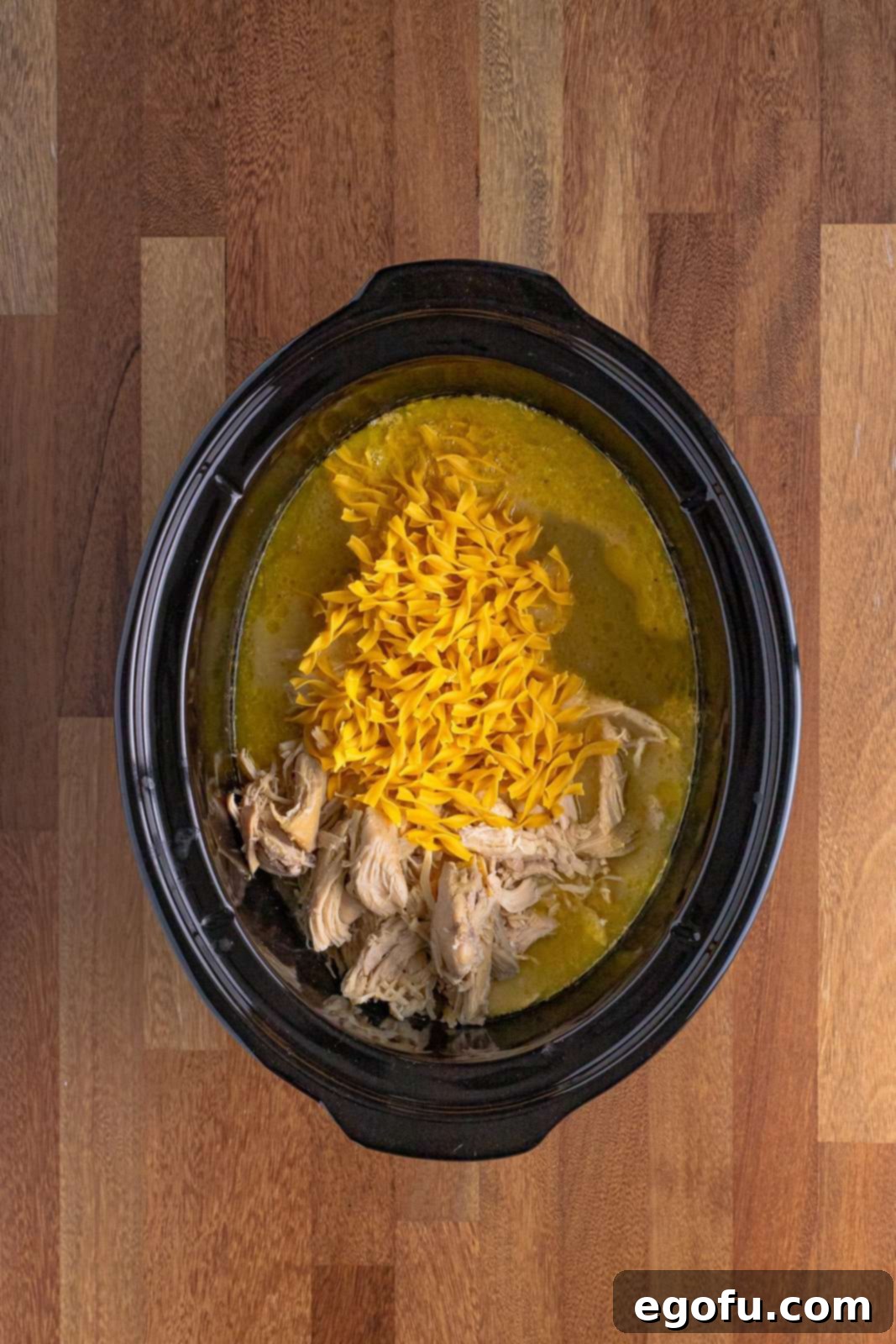 Shredded chicken and dry egg noodles added back into the slow cooker with the creamy broth, ready for the final cooking stage.