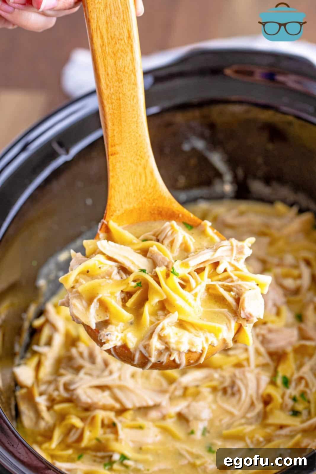 A wooden spoon scooping up a generous portion of creamy chicken and noodles directly from the slow cooker, showing the rich texture.
