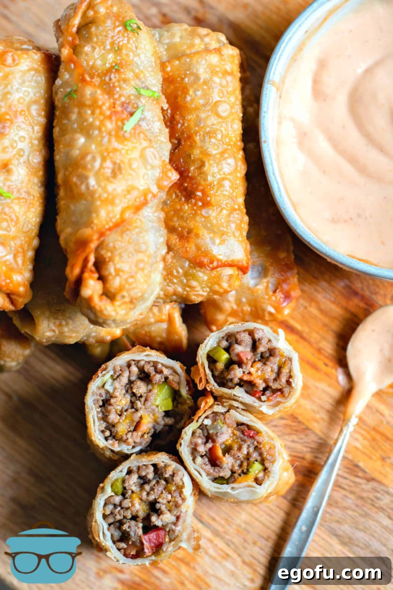 Crispy Cheeseburger Rolls 2 Fully cooked cheeseburger egg rolls, sliced and arranged on a wooden plate with burger dipping sauce.