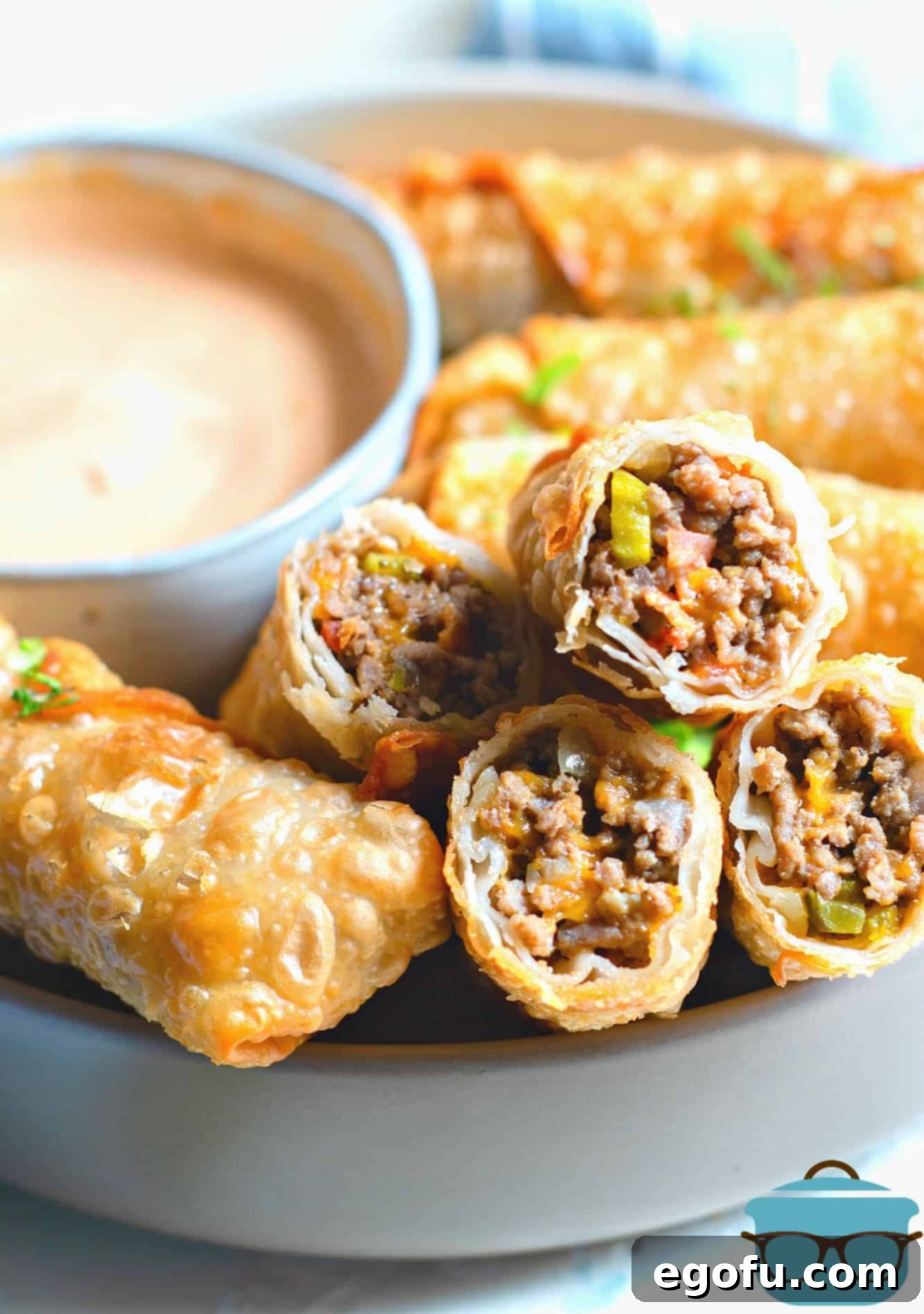 Crispy Cheeseburger Rolls 11 Cheeseburger Egg Rolls, fully cooked and arranged on a platter with the creamy dipping sauce in a small bowl in the background.