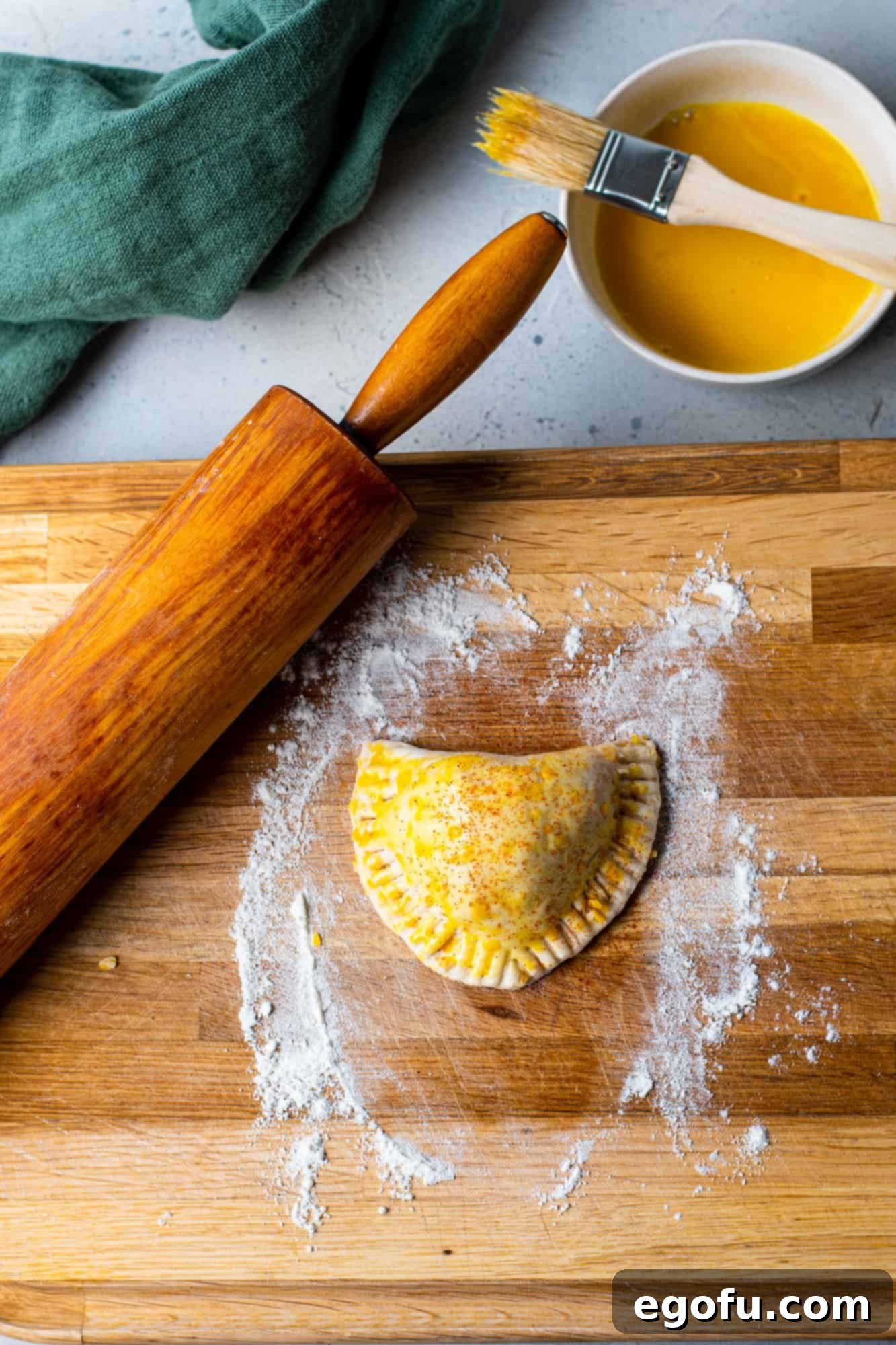 Egg wash and paprika on folded biscuit meat pies on a wooden cutting board, ready for baking.