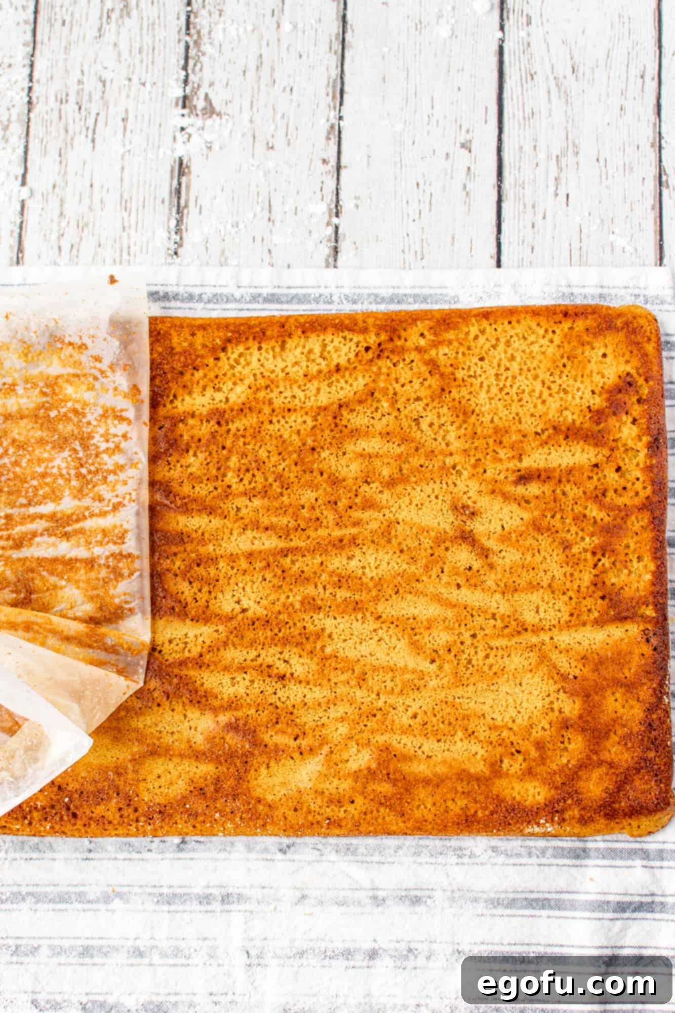 Parchment paper being gently peeled away from the warm, unrolled pumpkin cake, revealing its moist surface.