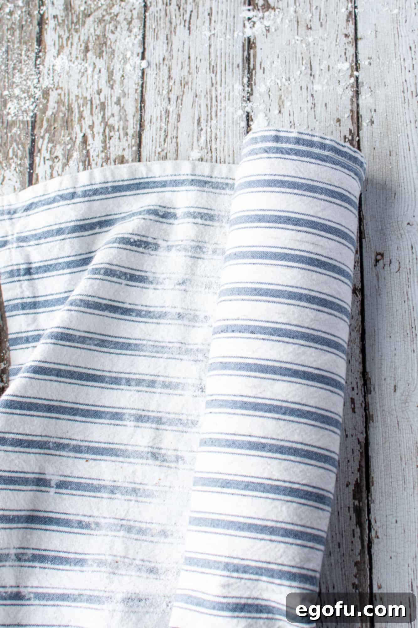 The warm pumpkin cake being rolled up from the short end with the powdered sugar-dusted kitchen towel, demonstrating the crucial rolling technique.