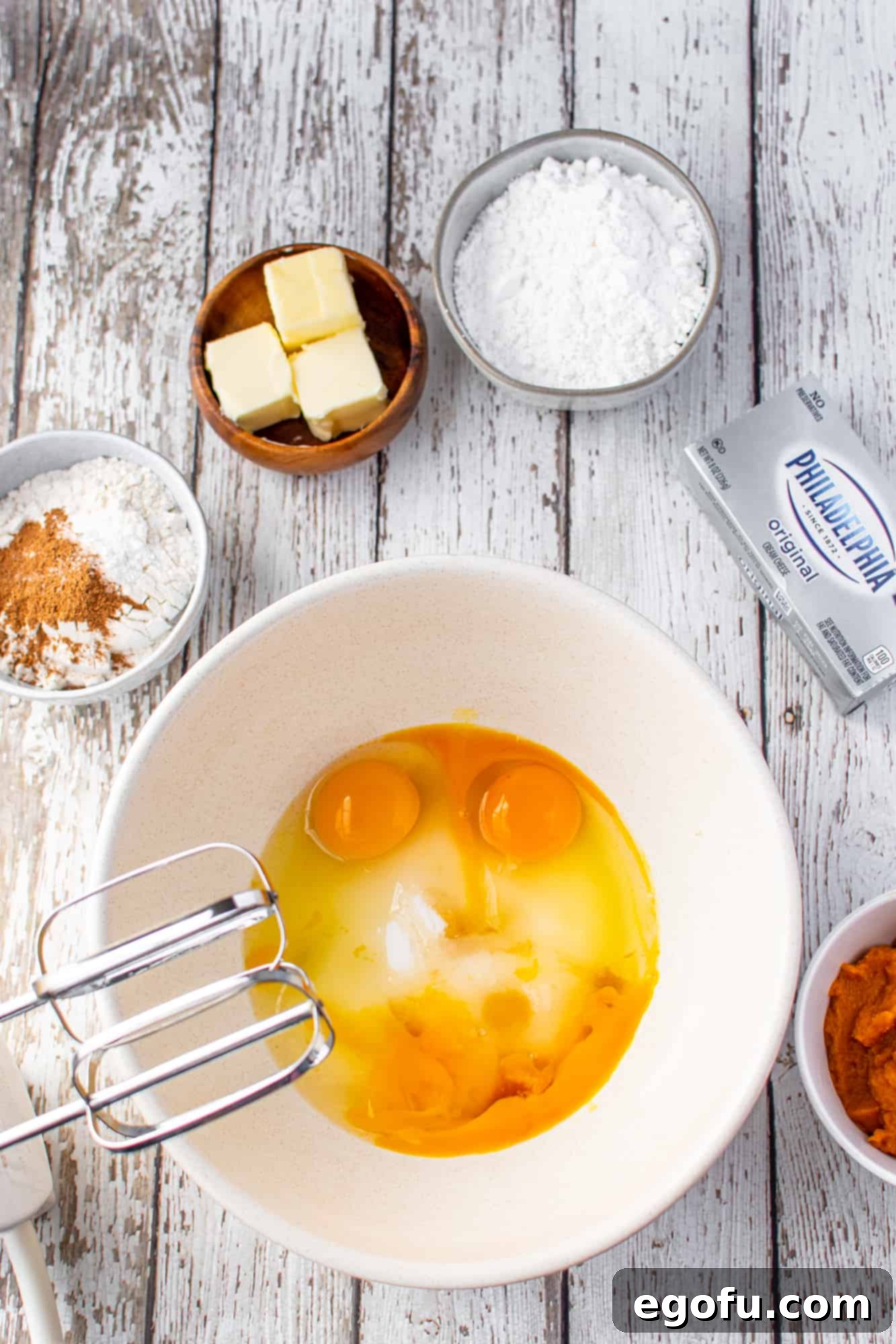 Granulated sugar and eggs being beaten together in a white mixing bowl with an electric mixer.