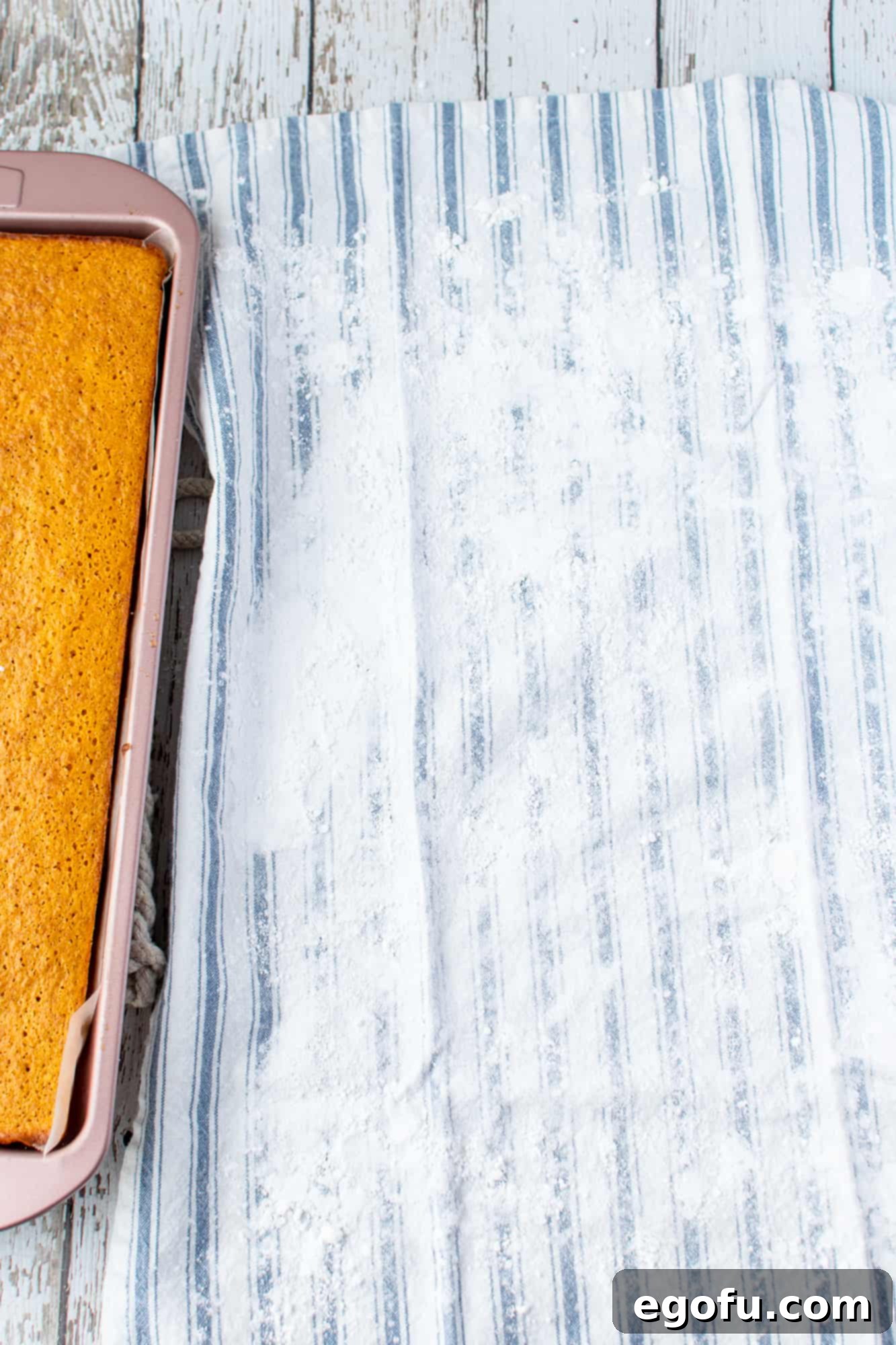 A clean kitchen towel lightly dusted with powdered sugar, ready to receive the warm pumpkin cake for rolling.
