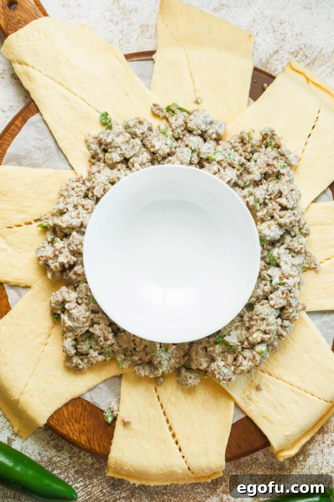 Sausage and cream cheese mixture carefully scooped onto the wide ends of crescent roll dough rectangles, arranged in a circular pattern around a central bowl on parchment paper.