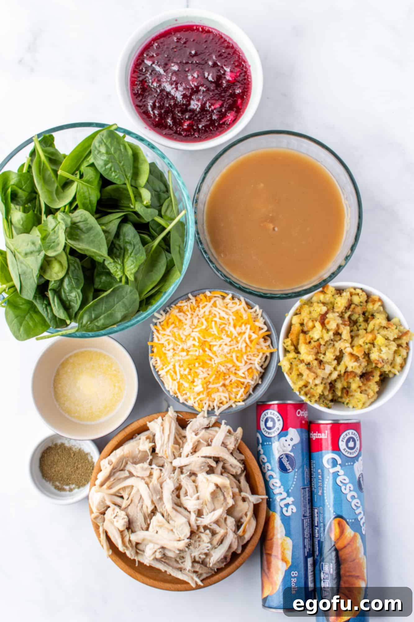 Festive Turkey Stuffing Crescent Bake 4 A flat lay photograph displaying all the ingredients needed for the Turkey and Stuffing Crescent Ring: crescent rolls, shredded turkey, stuffing, spinach leaves, cranberry sauce, Monterey Jack cheese, melted butter, poultry seasoning, and a bowl of turkey gravy.