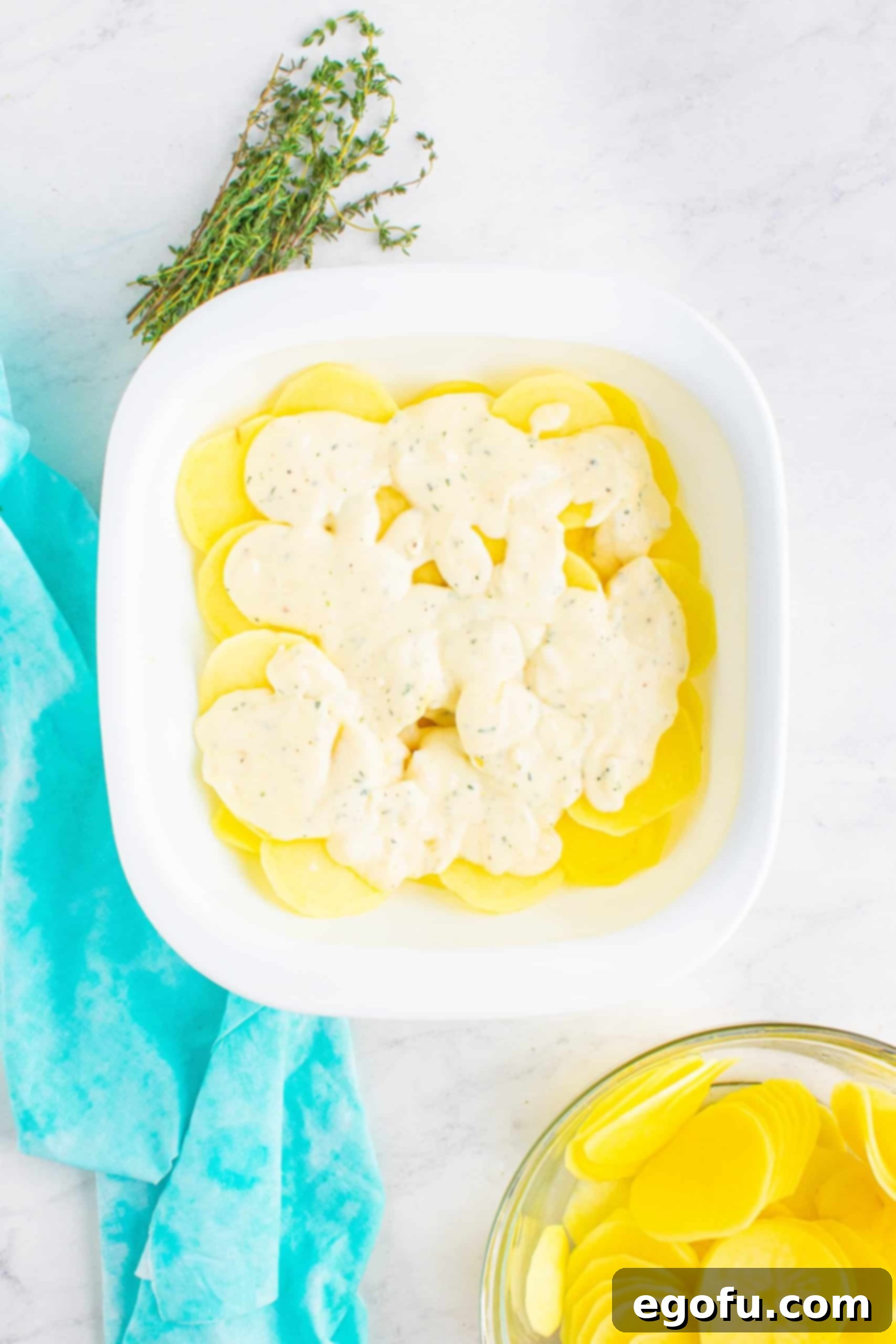 cheese sauce spread on top of sliced potatoes in a white, square baking dish.