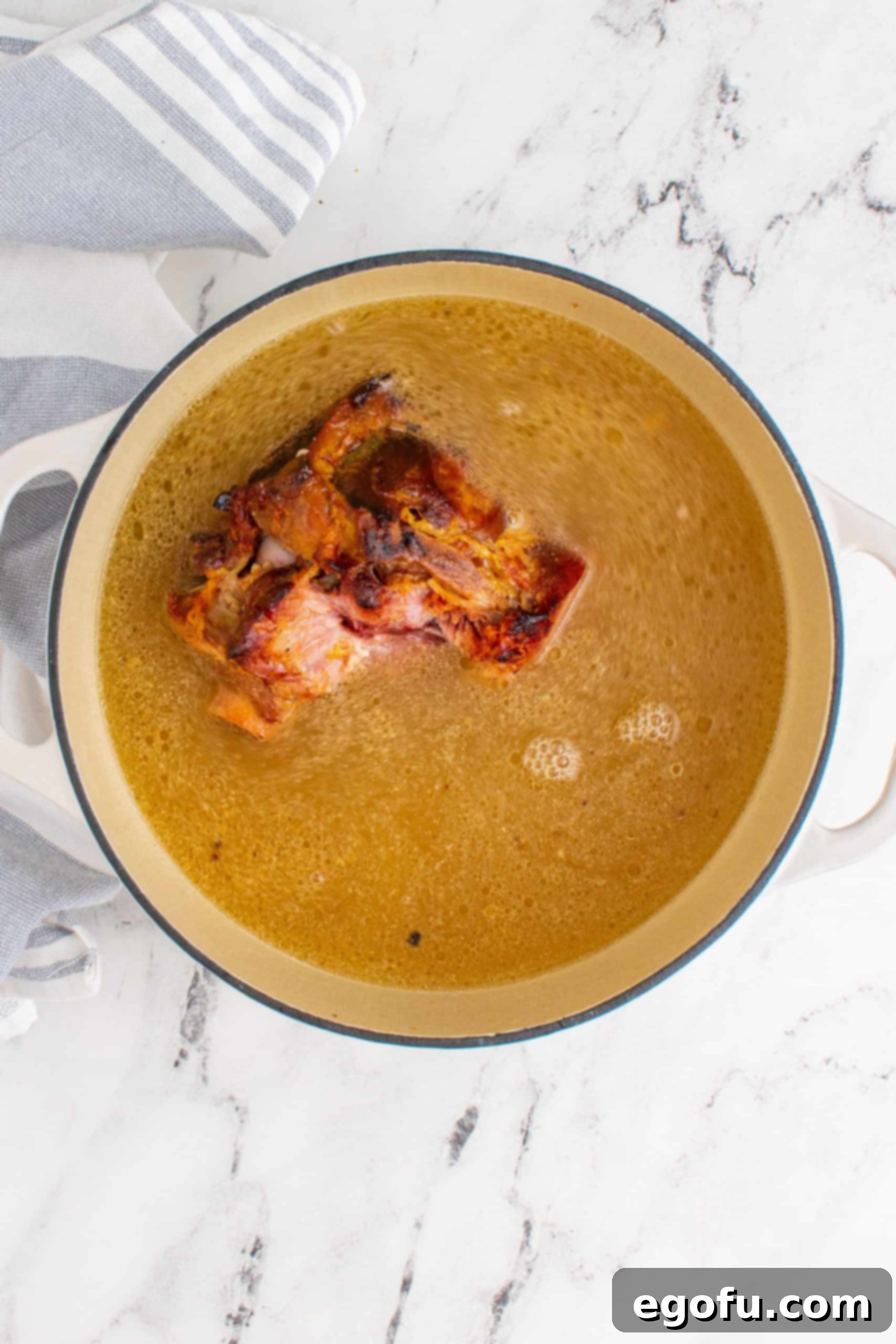 Chicken stock and ham bone simmering in a white pot on a marble surface, infusing the broth with flavor.
