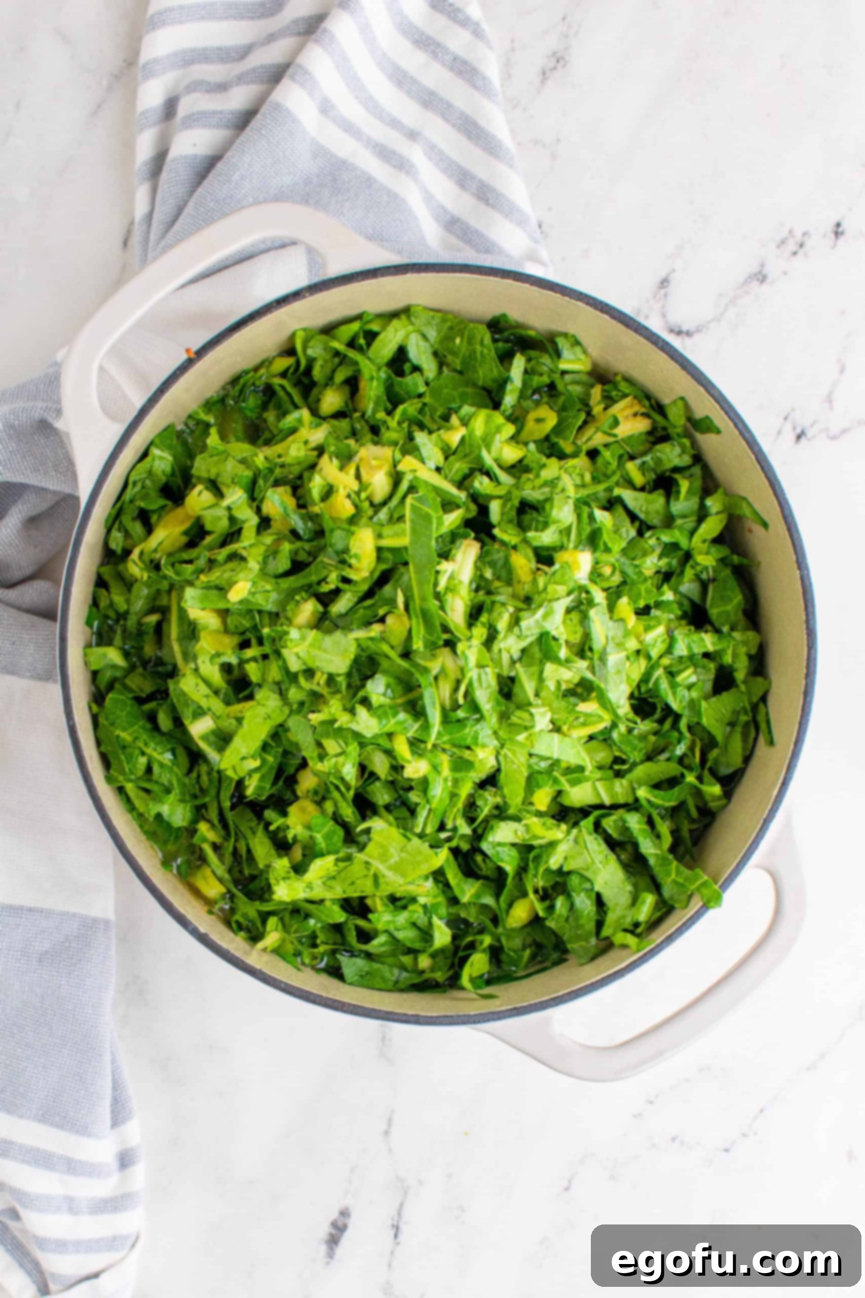 Shredded collard greens being added to a white Dutch oven pot on a marble surface, ready to be wilted.