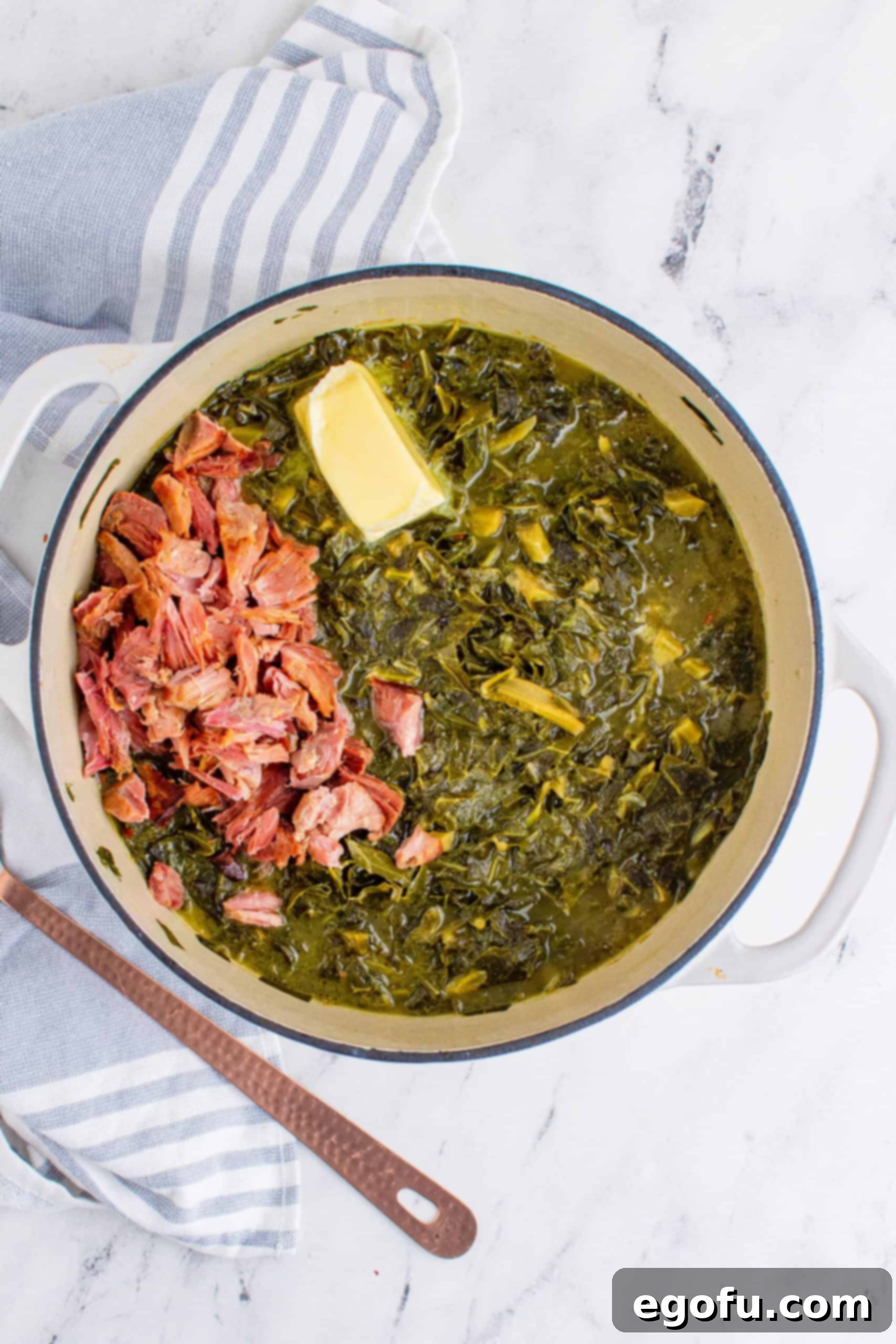 Shredded ham added to cooked collard greens with butter melting in a white pot, alongside a copper spoon.