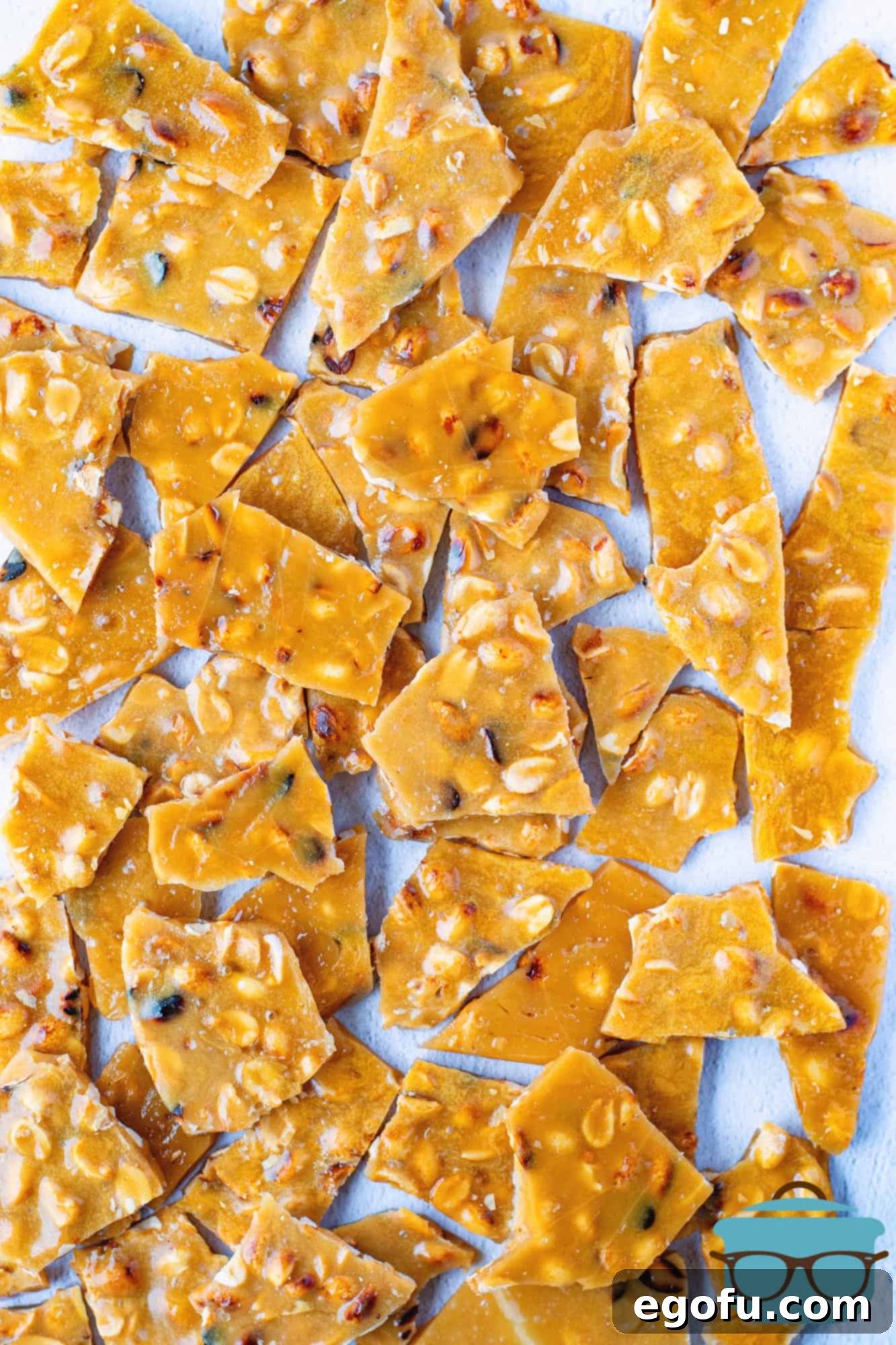 Golden Crunch Peanut Brittle 10 Peanut brittle broken into numerous glistening, golden pieces, scattered on a sheet of white parchment paper, ready to be enjoyed.