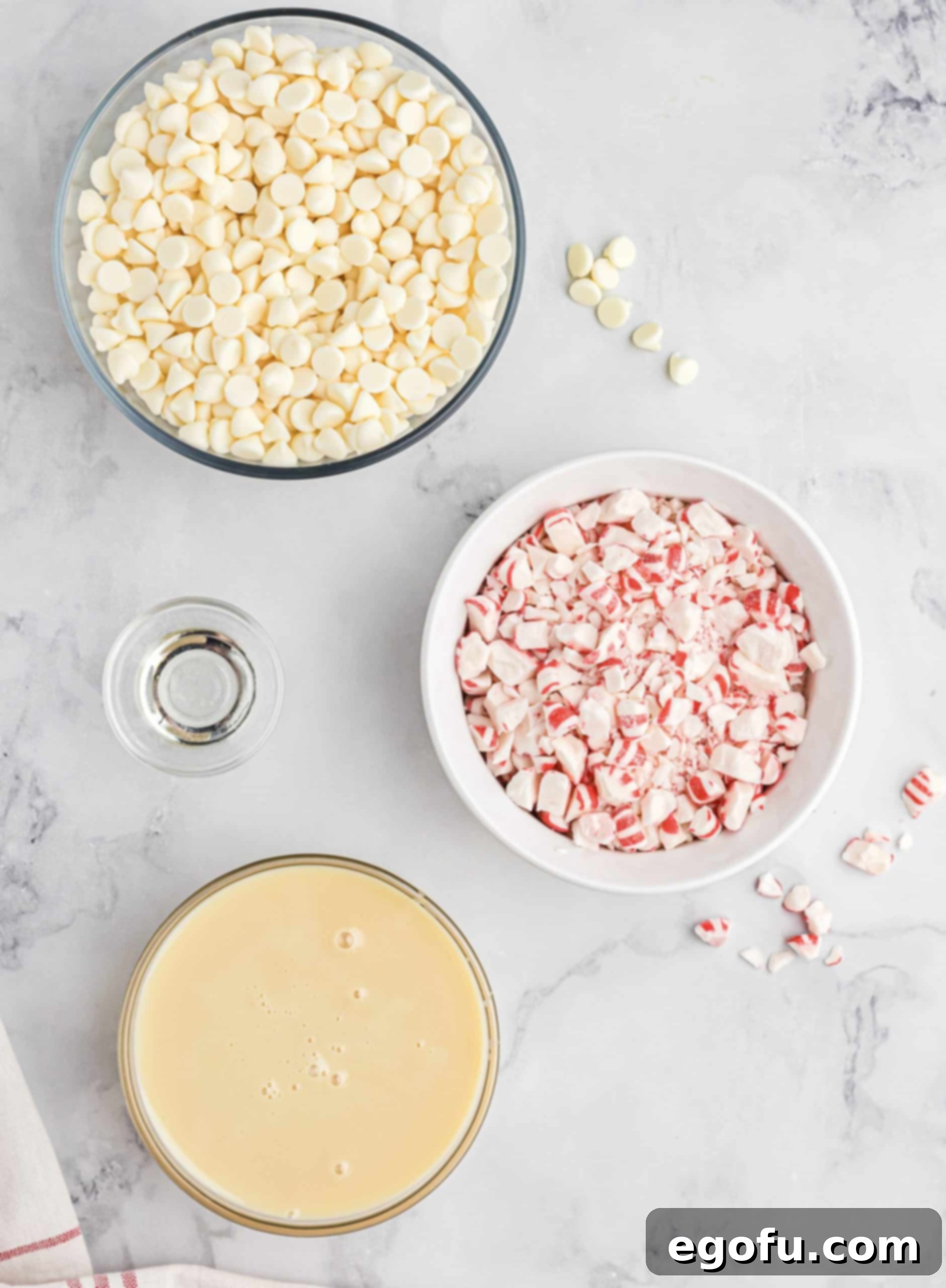 A beautiful flat lay photograph displaying the key ingredients for Candy Cane Fudge: a bowl of 3 cups of white chocolate chips (approximately 1 lb), a can of 14 oz sweetened condensed milk, a small bottle of 1 tsp peppermint extract, and a bowl of 2 cups of vibrant crushed candy canes or peppermint candies.