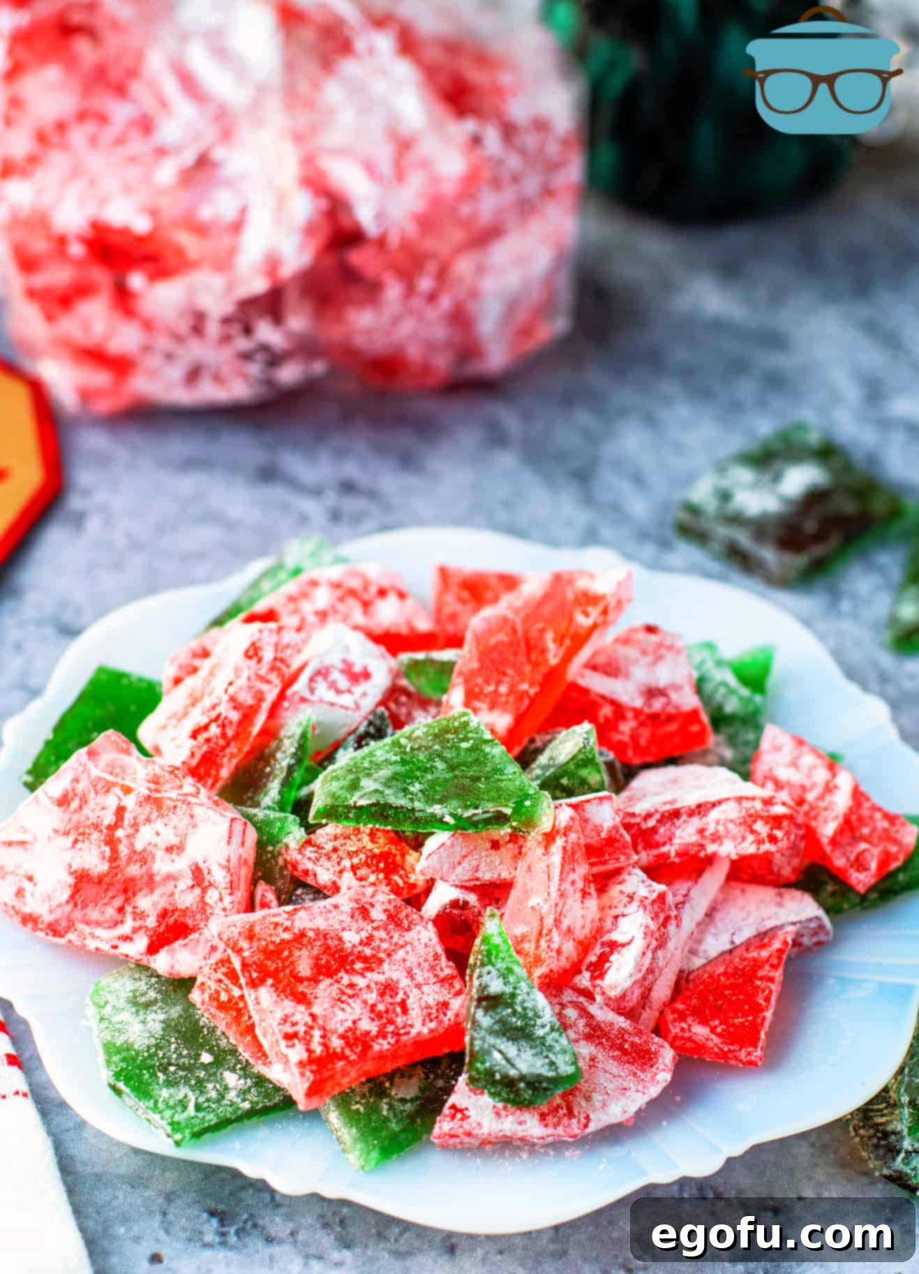 Pieces of red and green Apple Cinnamon Rock Candy displayed on a white plate, with additional red candy in a bag in the blurred background.