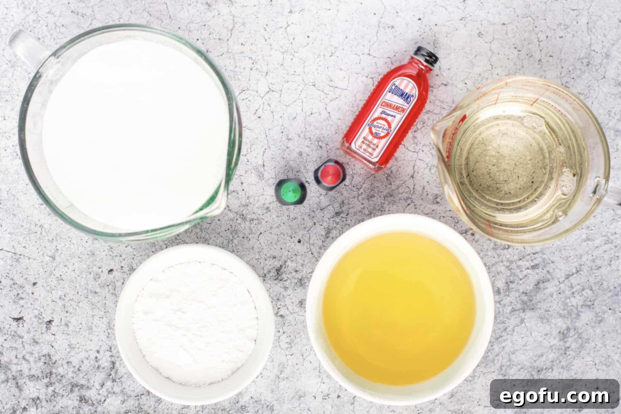 A flat lay of the essential ingredients for Apple Cinnamon Rock Candy: apple juice, granulated sugar, light corn syrup, vibrant red and green liquid food coloring, a bottle of cinnamon flavoring, and powdered sugar.