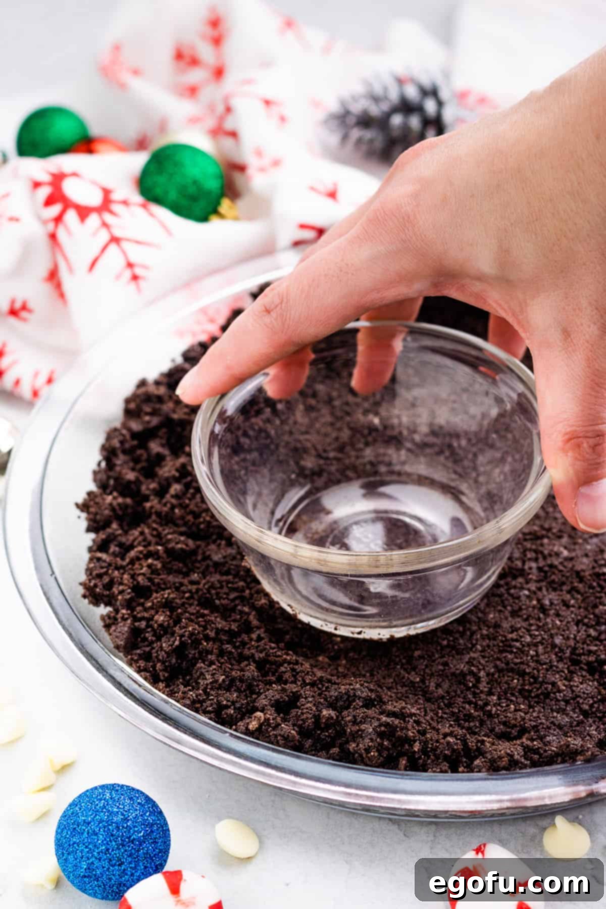 A hand using the bottom of a glass to firmly press the Oreo cookie crumb mixture into the base of a clear glass pie plate, forming the crust.