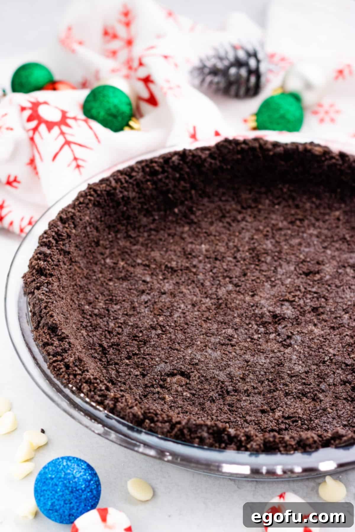 A freshly baked Oreo cookie pie crust in a clear round pie pan, set against a festive background with small Christmas decorations.