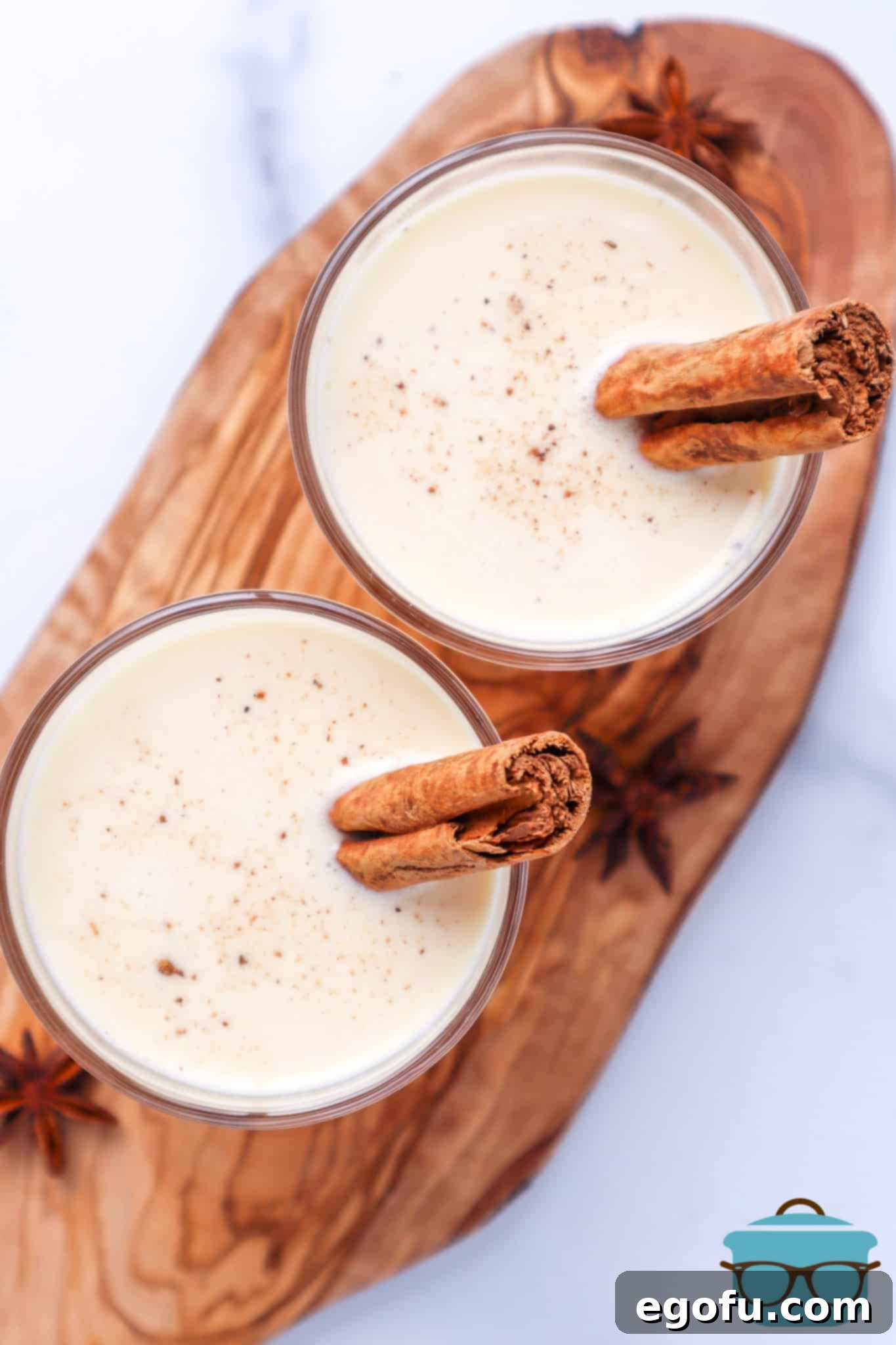 Instant Pot Egg Nog shown in two small, clear glasses with sticks of cinnamon in each glass shown on a wooden cutting board.