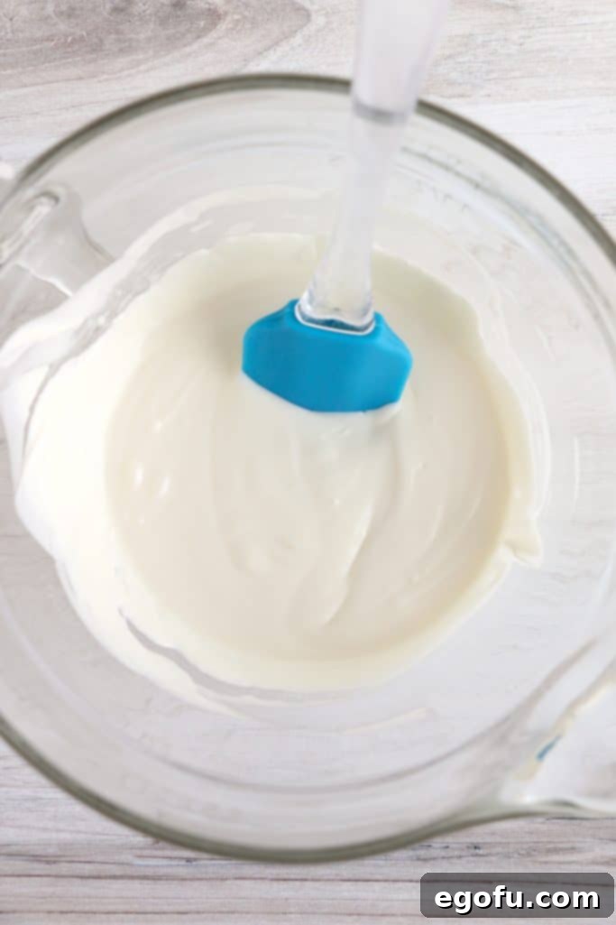 Perfectly melted vanilla candy coating in a clear bowl with a blue scraper, showing its smooth, creamy consistency.