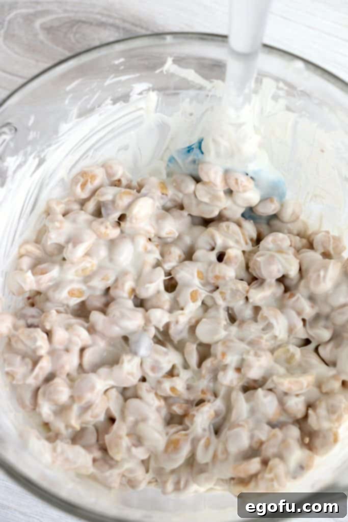 Salted peanuts being stirred into the melted vanilla almond bark in a clear bowl, ensuring thorough coating.