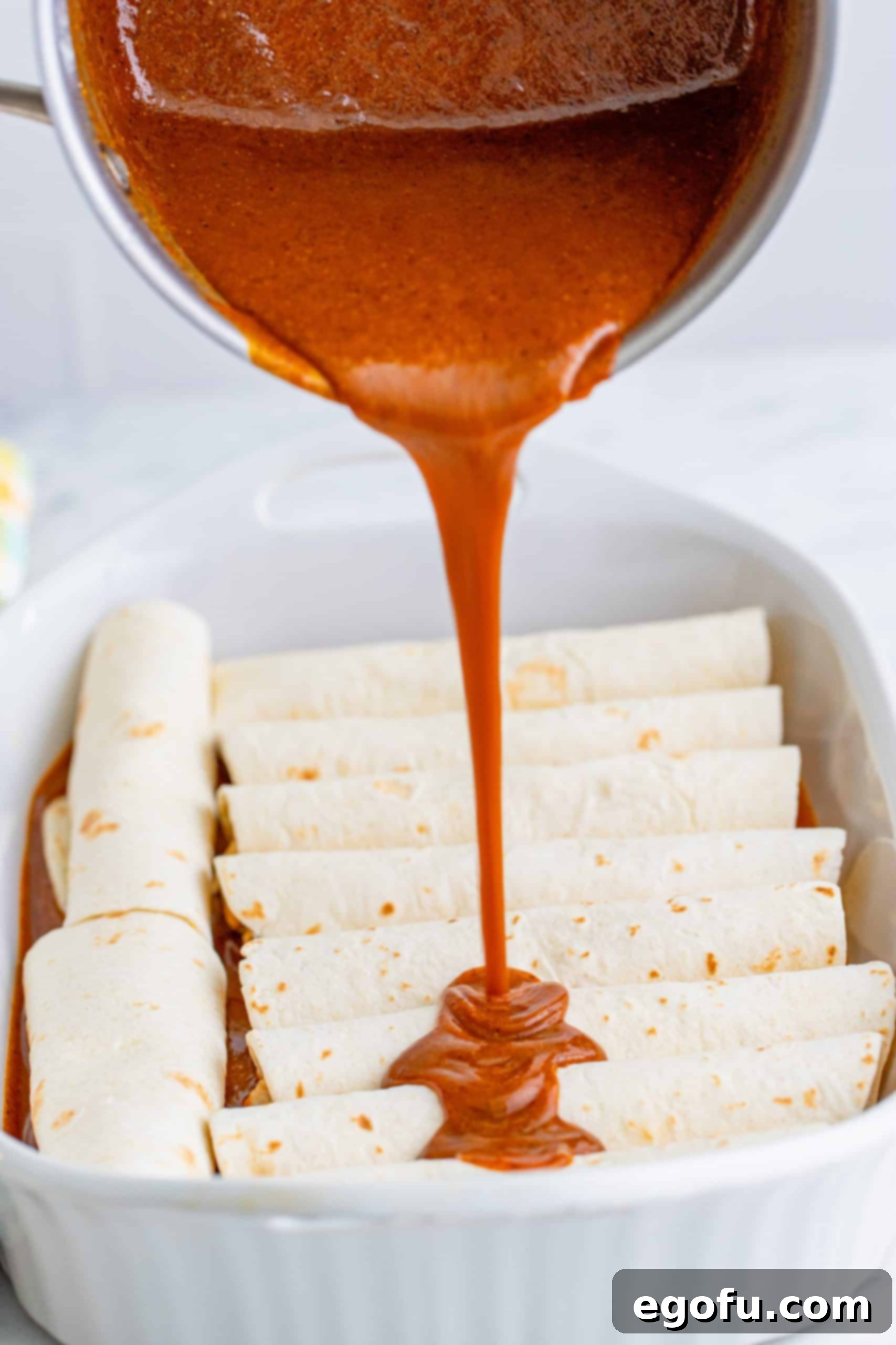 Pouring enchilada sauce on top of rolled flour chicken enchiladas in a baking dish, creating a luscious covering.