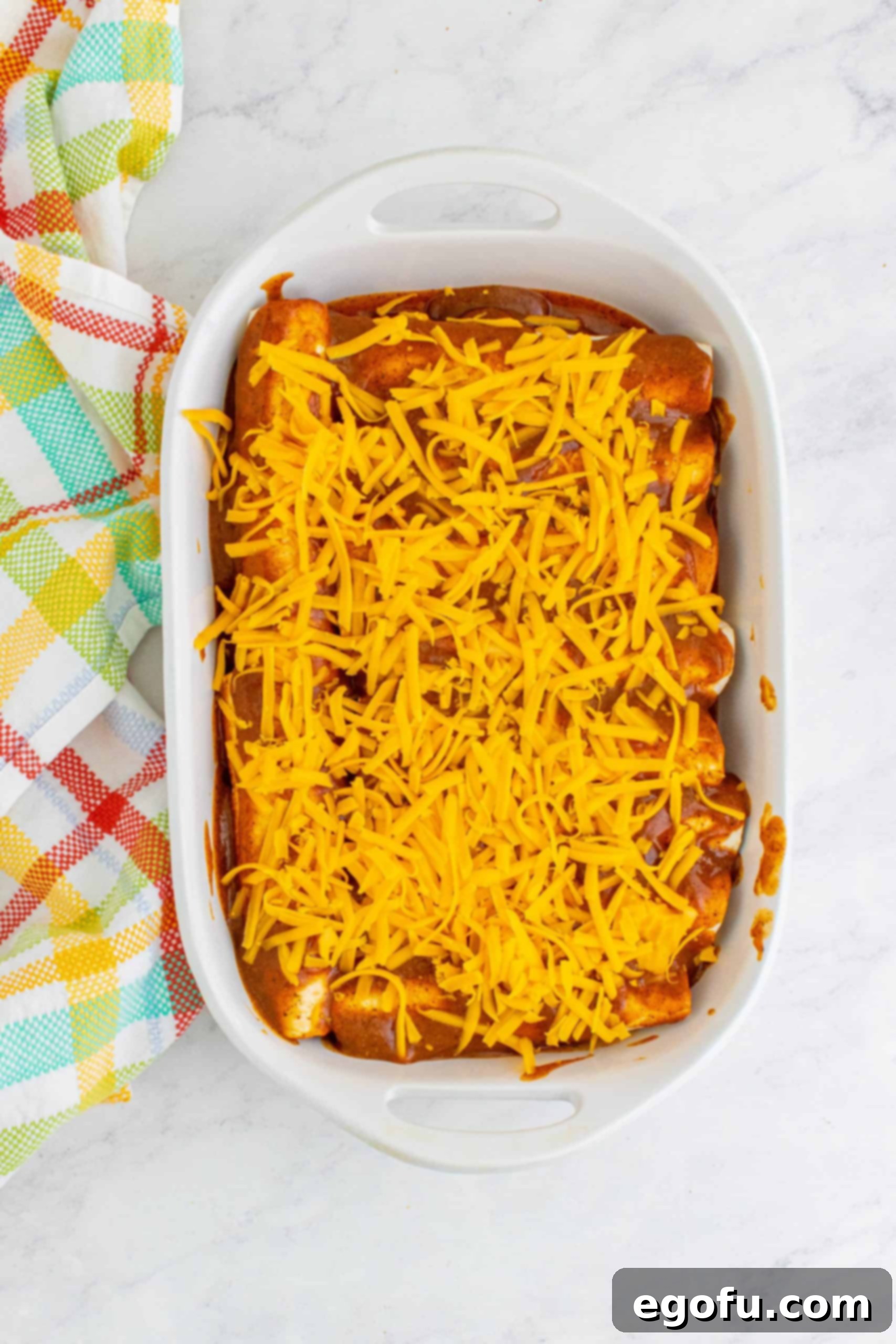 Shredded cheddar cheese sprinkled evenly on top of prepared enchiladas in baking dish, ready for baking.