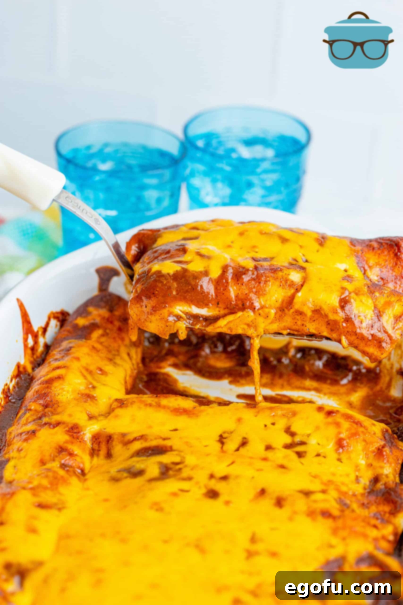 Scooping out two red sauce enchiladas out of a baking dish with a spatula, showcasing their golden-brown perfection.