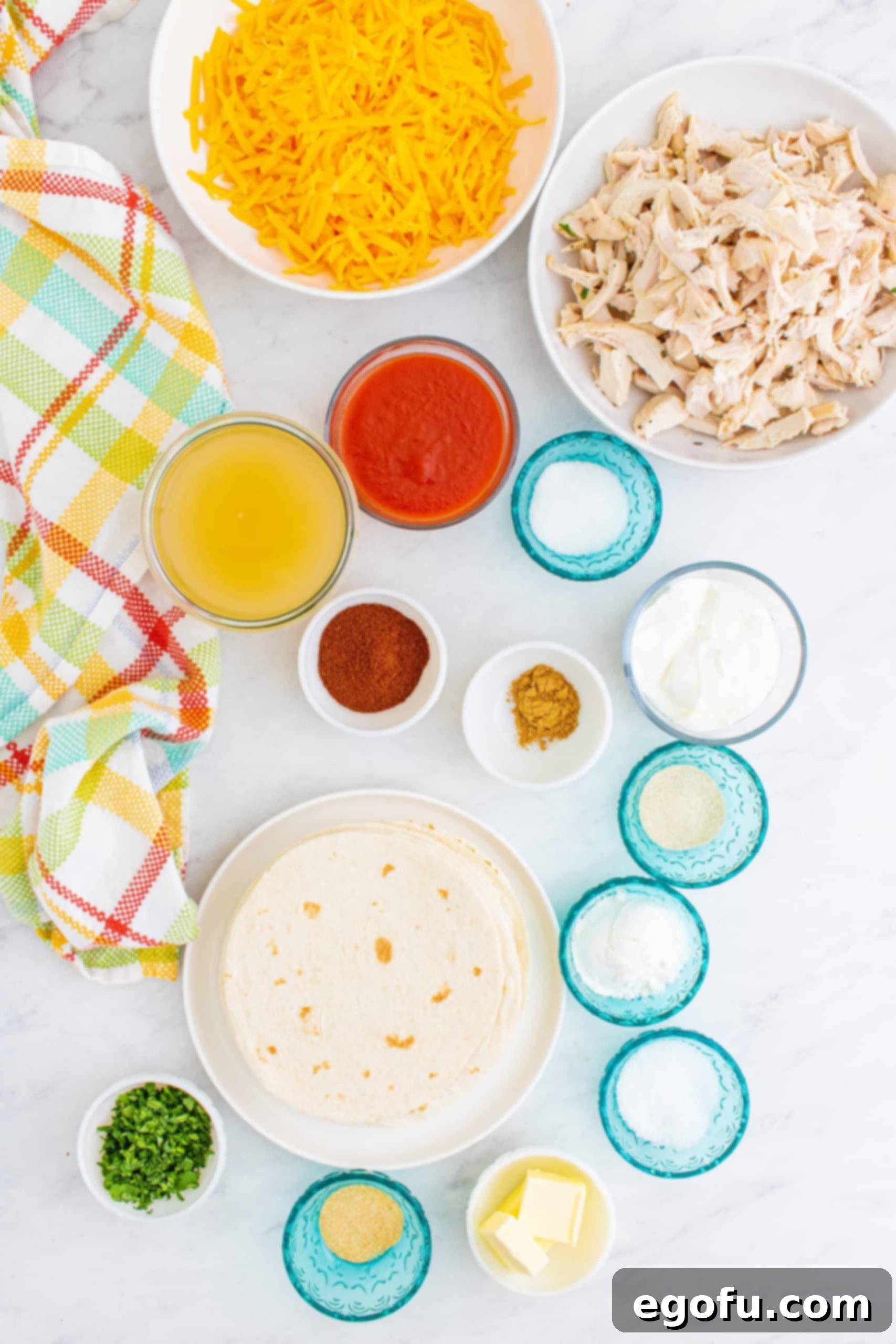 An array of ingredients laid out: butter, chili powder, garlic powder, onion powder, ground cumin, cornstarch, chicken stock, tomato sauce, granulated sugar, kosher salt, shredded cooked chicken, shredded cheddar cheese, sour cream, fresh cilantro, and flour tortillas.