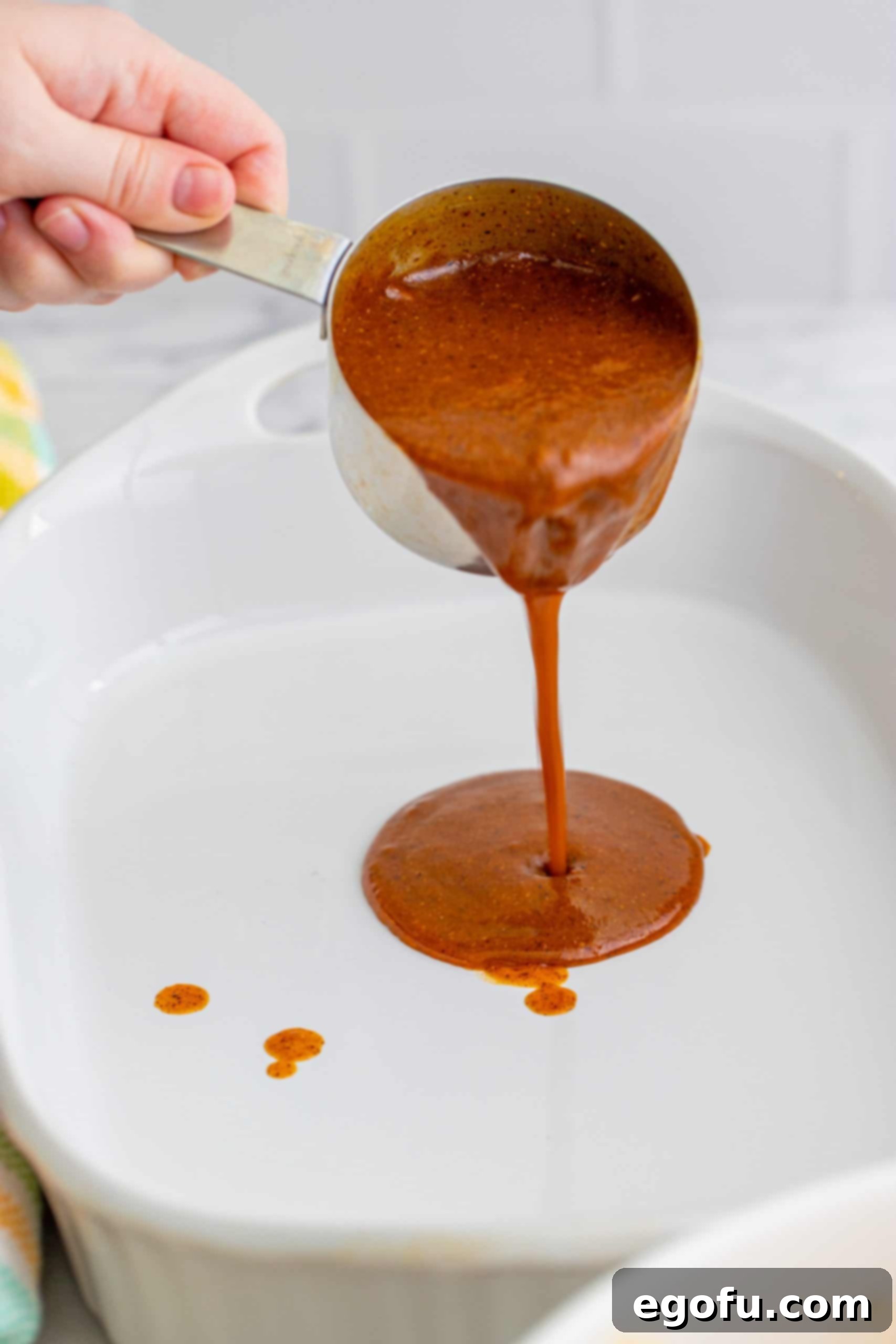 Pouring homemade red sauce from a cup into a white baking dish, ensuring an even layer.