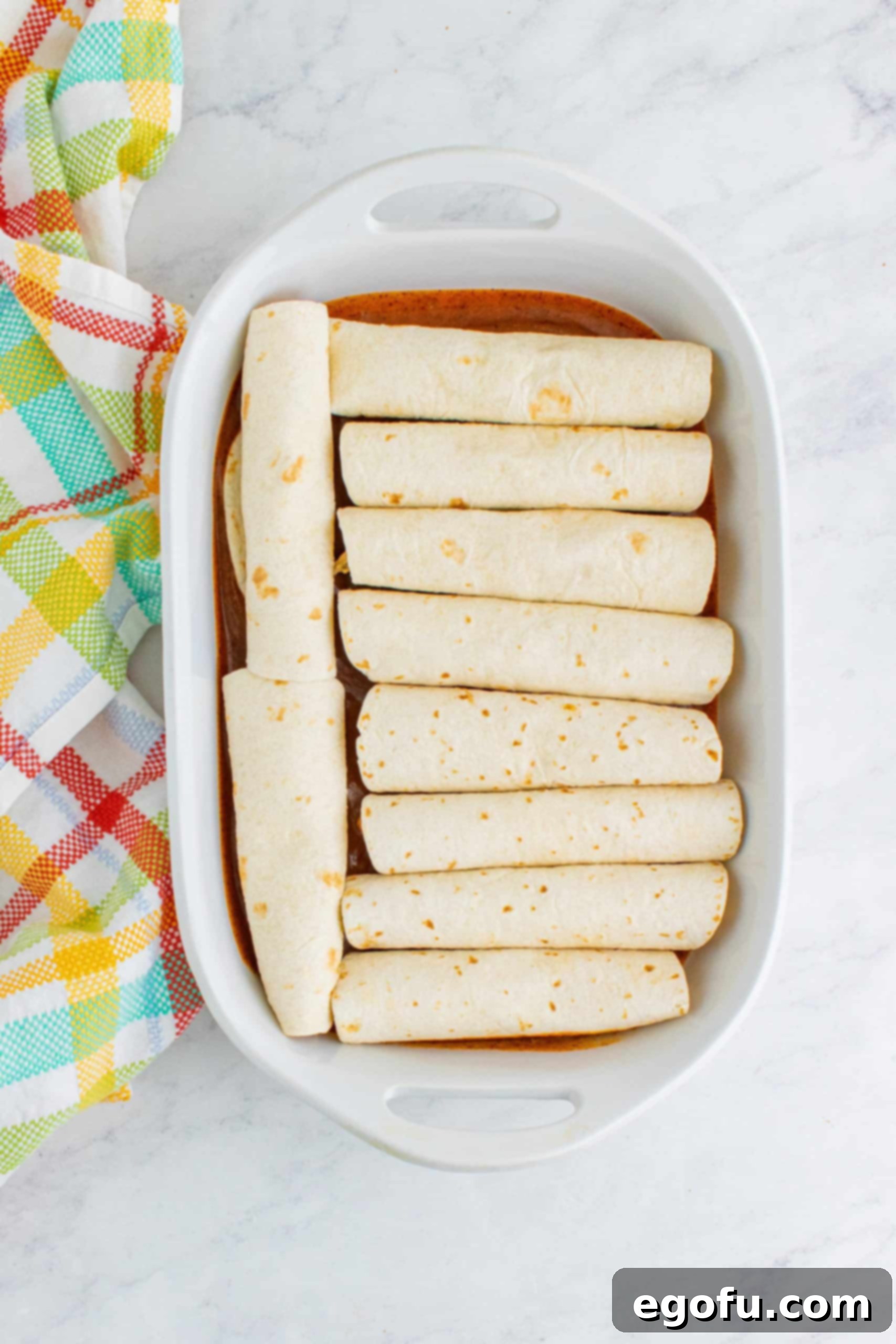 10 rolled tortillas placed in a single layer in a white baking dish, ready for more sauce and cheese.