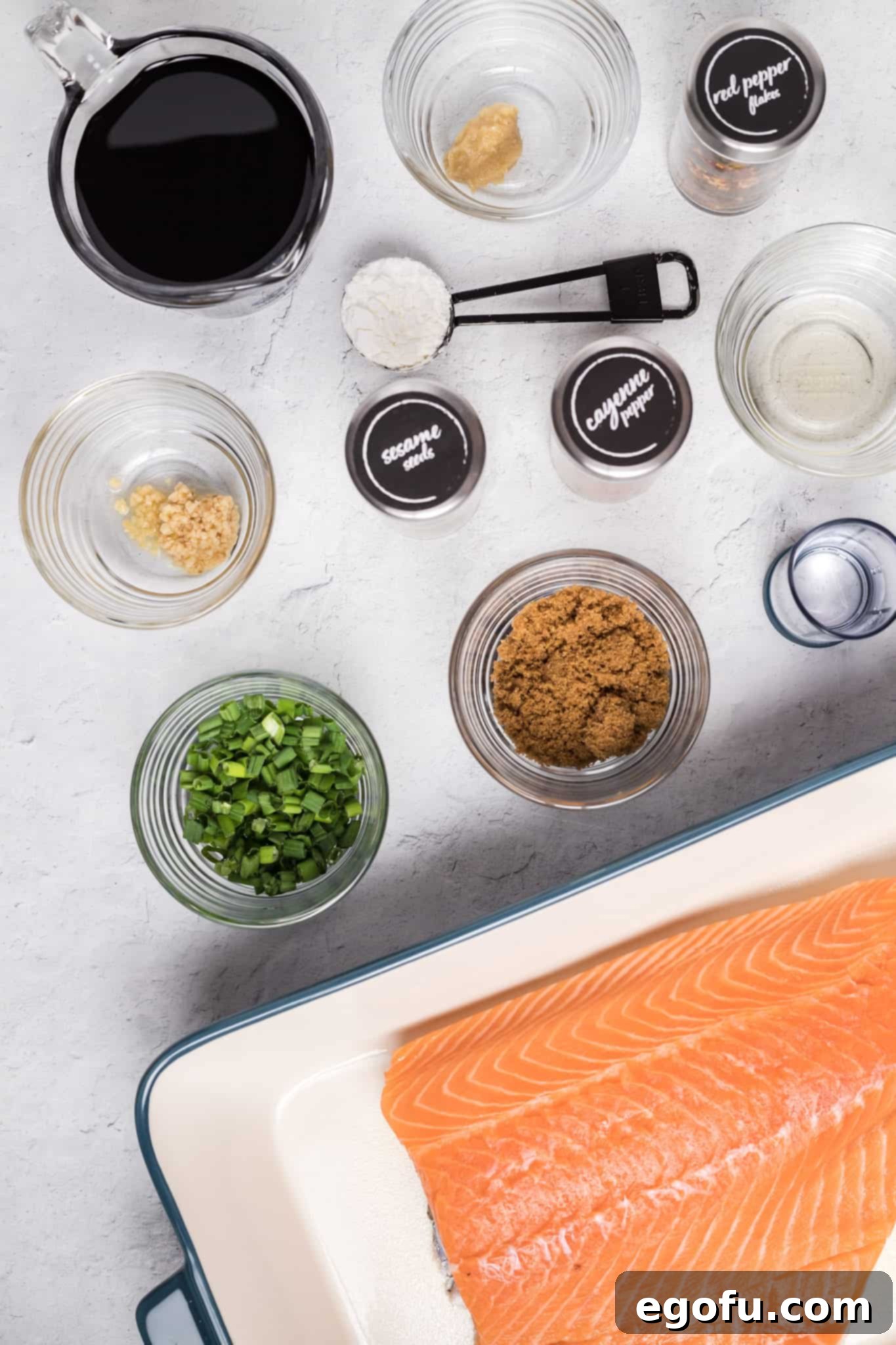 A vibrant flat lay showcasing all the fresh ingredients needed to make homemade teriyaki salmon: center cut salmon fillet, bottle of soy sauce, minced ginger, minced garlic, cornstarch, water, brown sugar, rice vinegar, sesame seeds, cayenne pepper, and a bunch of green onions.