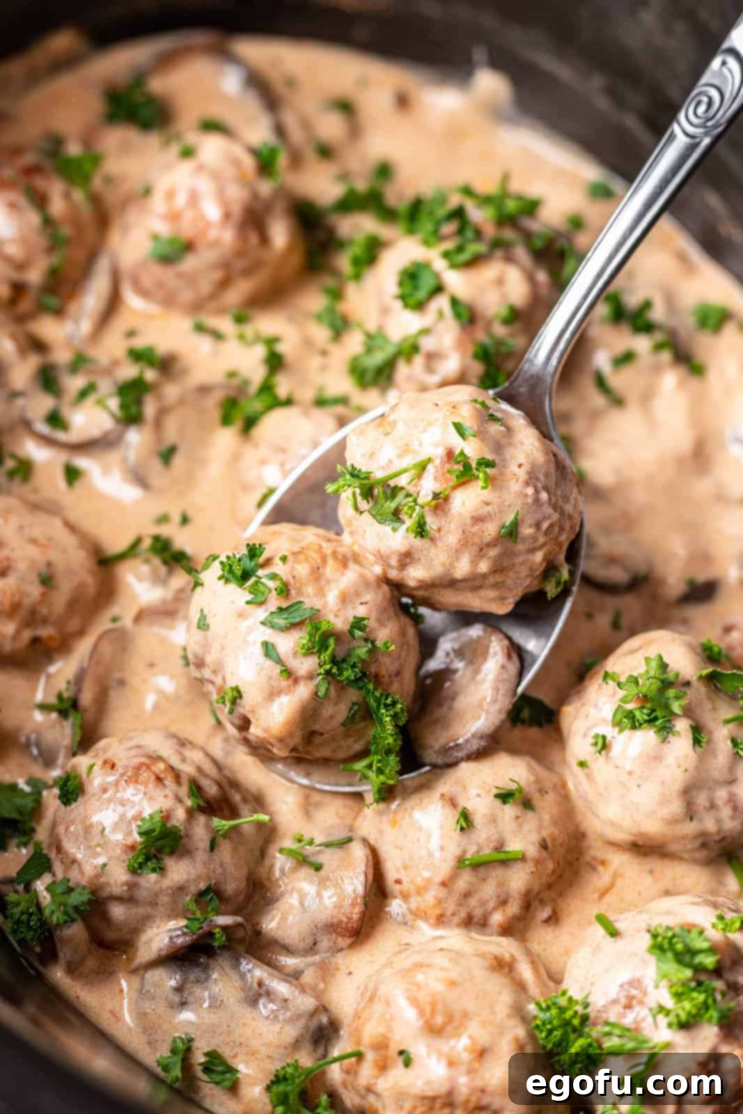 A spoon lifting two meatballs over the thick and creamy meatball stroganoff sauce in an oval slow cooker.