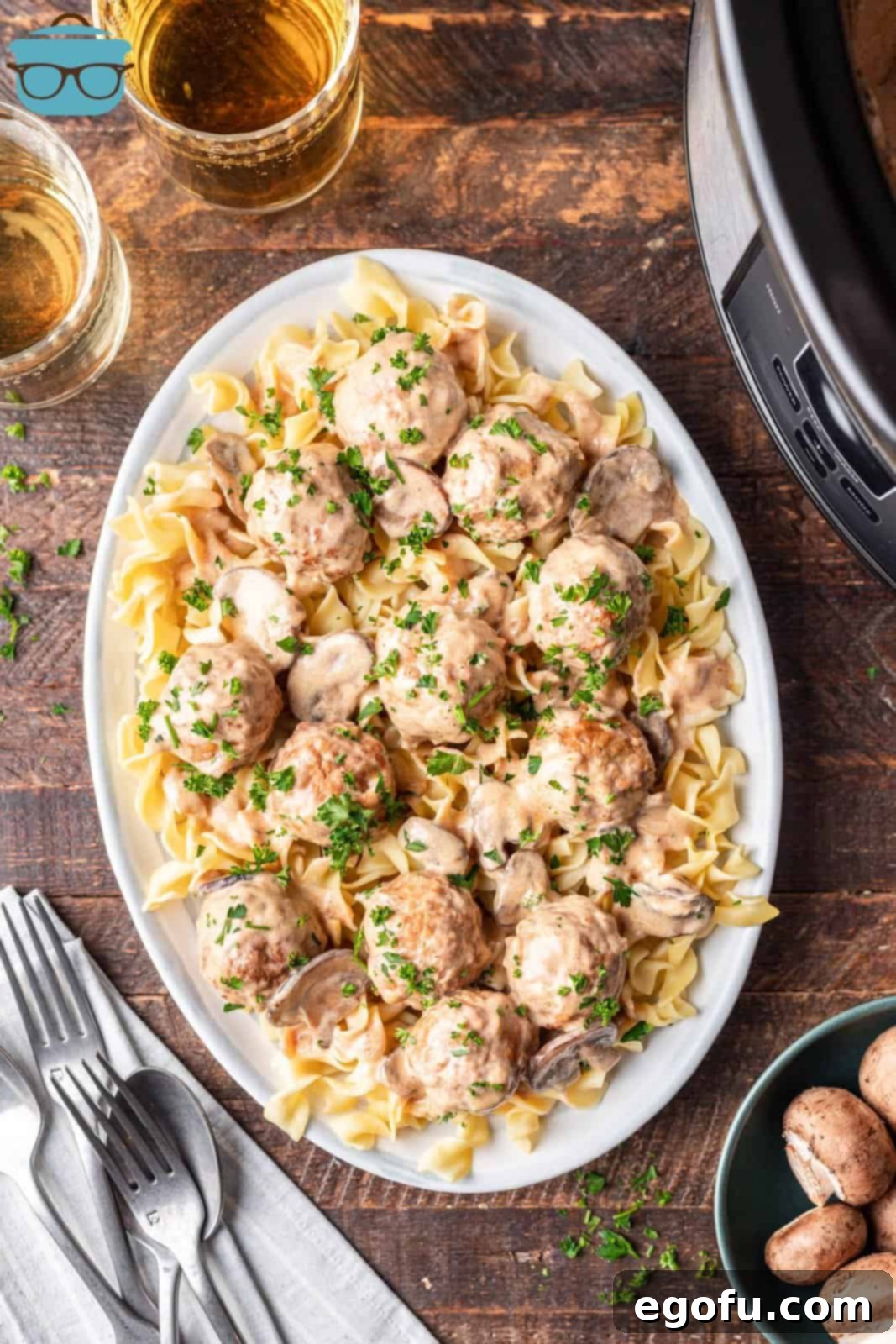 An overhead photo of egg noodles topped with Crock Pot Meatball Stroganoff on a large oval plate, placed on a wooden surface with the slow cooker nearby.