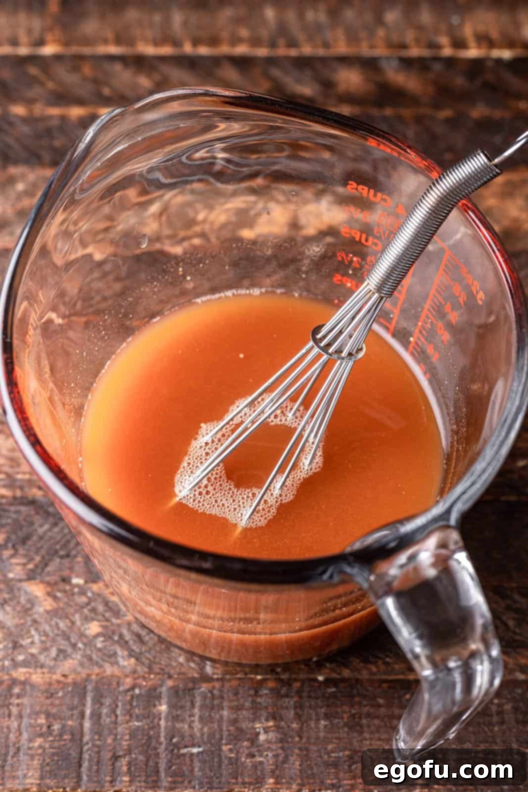 Beef broth, tomato paste, and mustard mixed together with a whisk in a clear measuring cup.