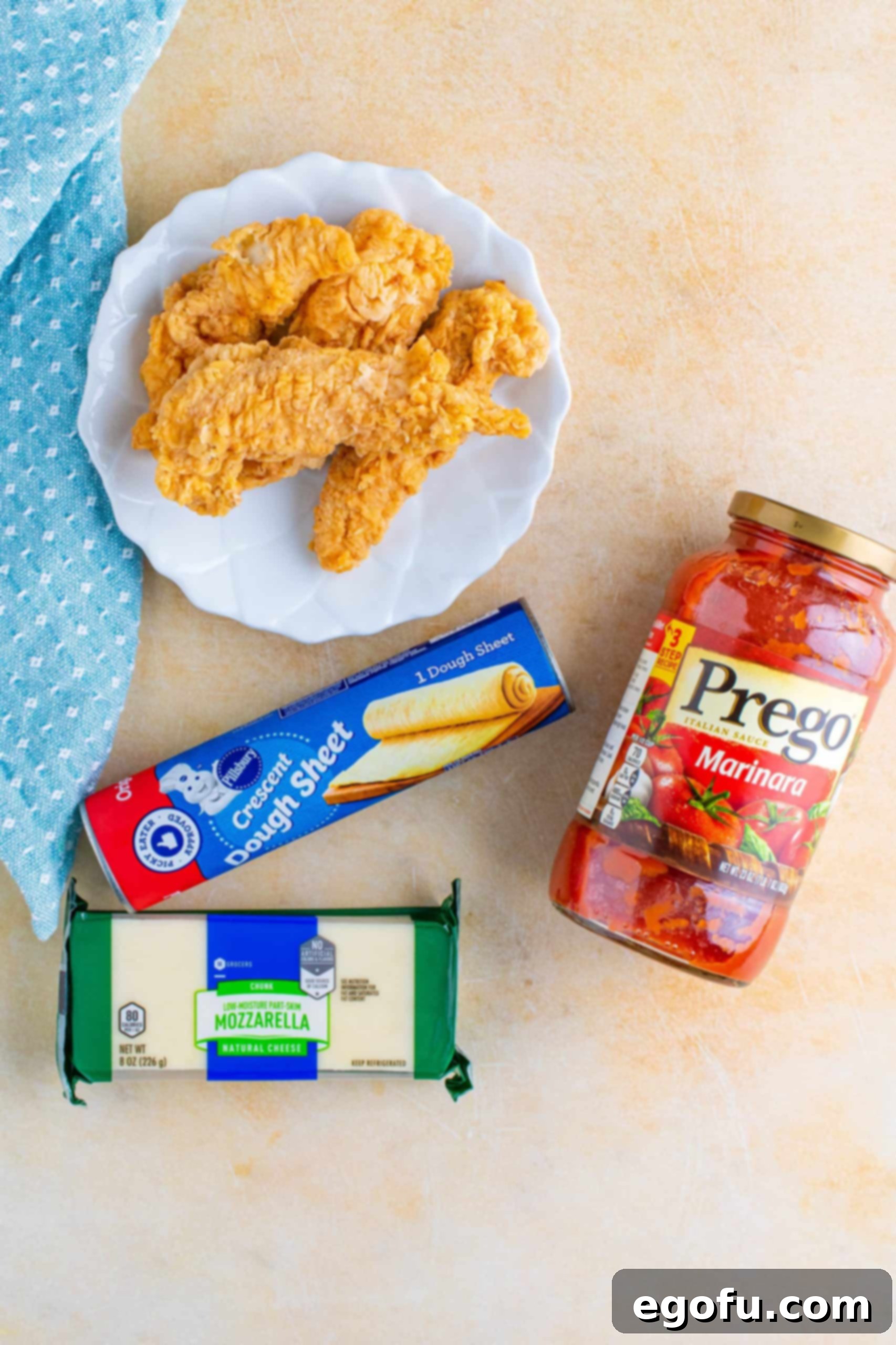 A flat lay of ingredients for chicken parmesan cups, including a can of crescent dough, shredded mozzarella, fried chicken tenders, and marinara sauce.